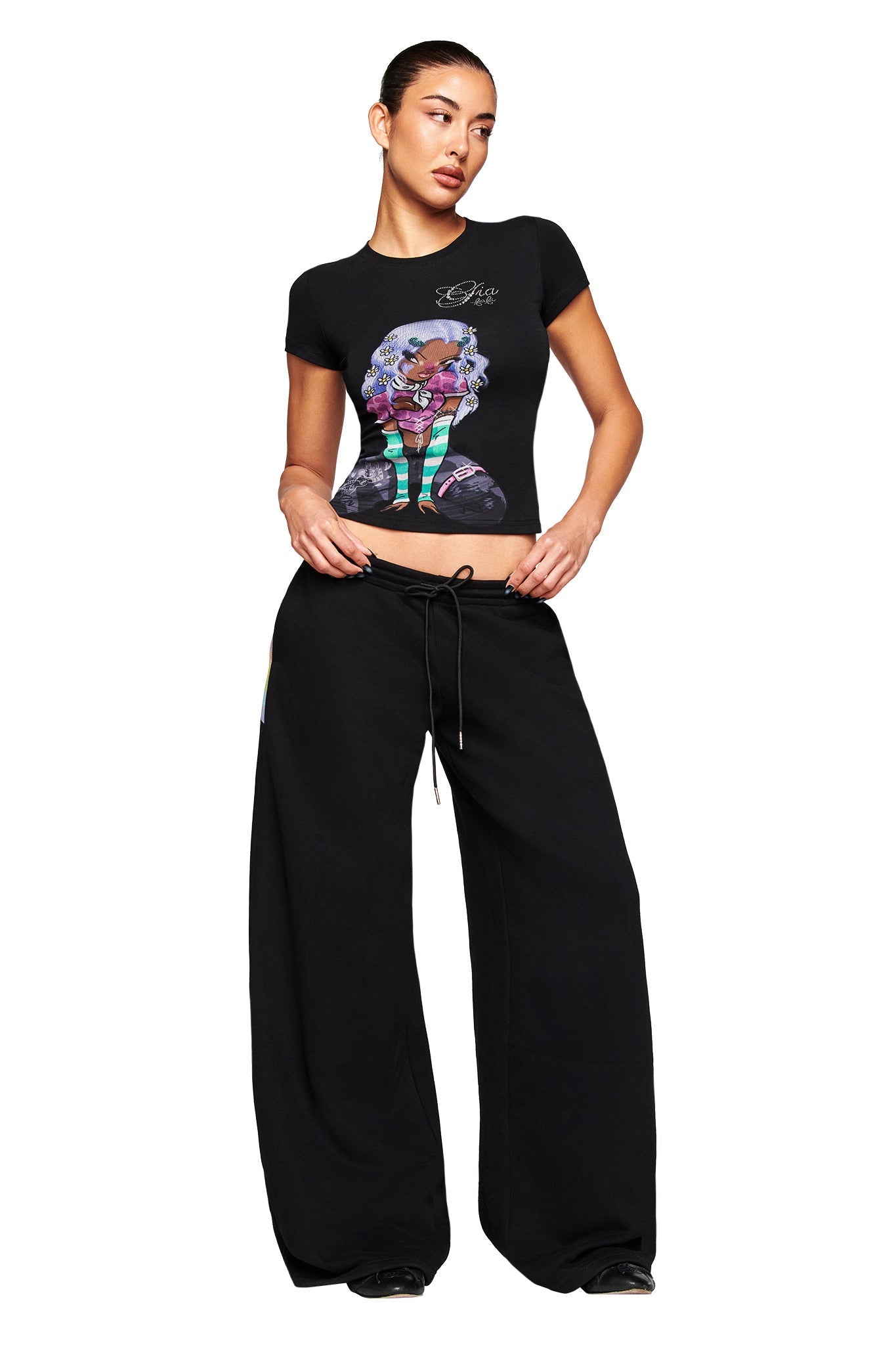 A woman stands against a white background wearing the I.AM.GIA FAFI T-SHIRT in black, featuring a colorful cartoon graphic, paired with loose black drawstring pants. She poses with her hands on her pants and looks to the side.