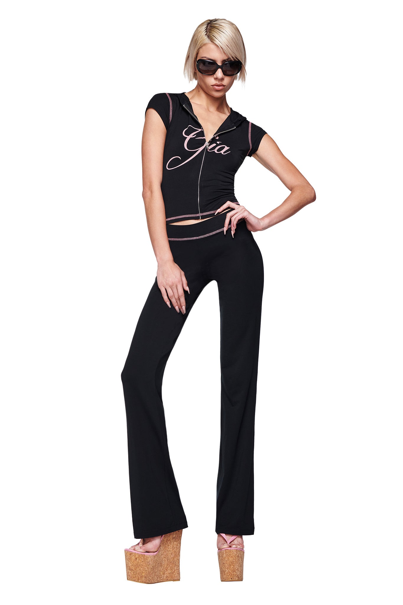 A woman in sunglasses strikes a confident pose against a white background, wearing the I.AM.GIA BLARE CAP SLEEVE HOODIE in black with matching pants and tall cork platform sandals.