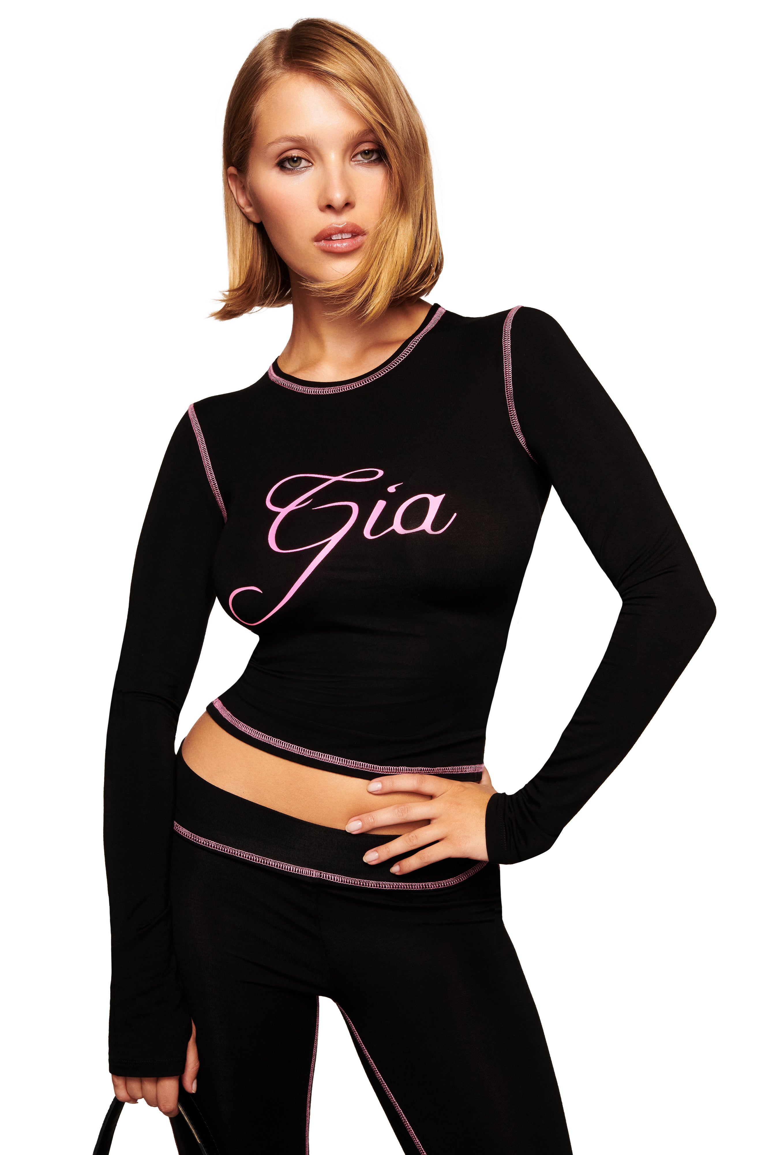 A woman with a sleek blond bob poses confidently in the I.AM.GIA BLARE LONG SLEEVE TOP - BLACK, featuring pink trim and “Gia” cursive. She holds a black handbag and looks directly at the camera.