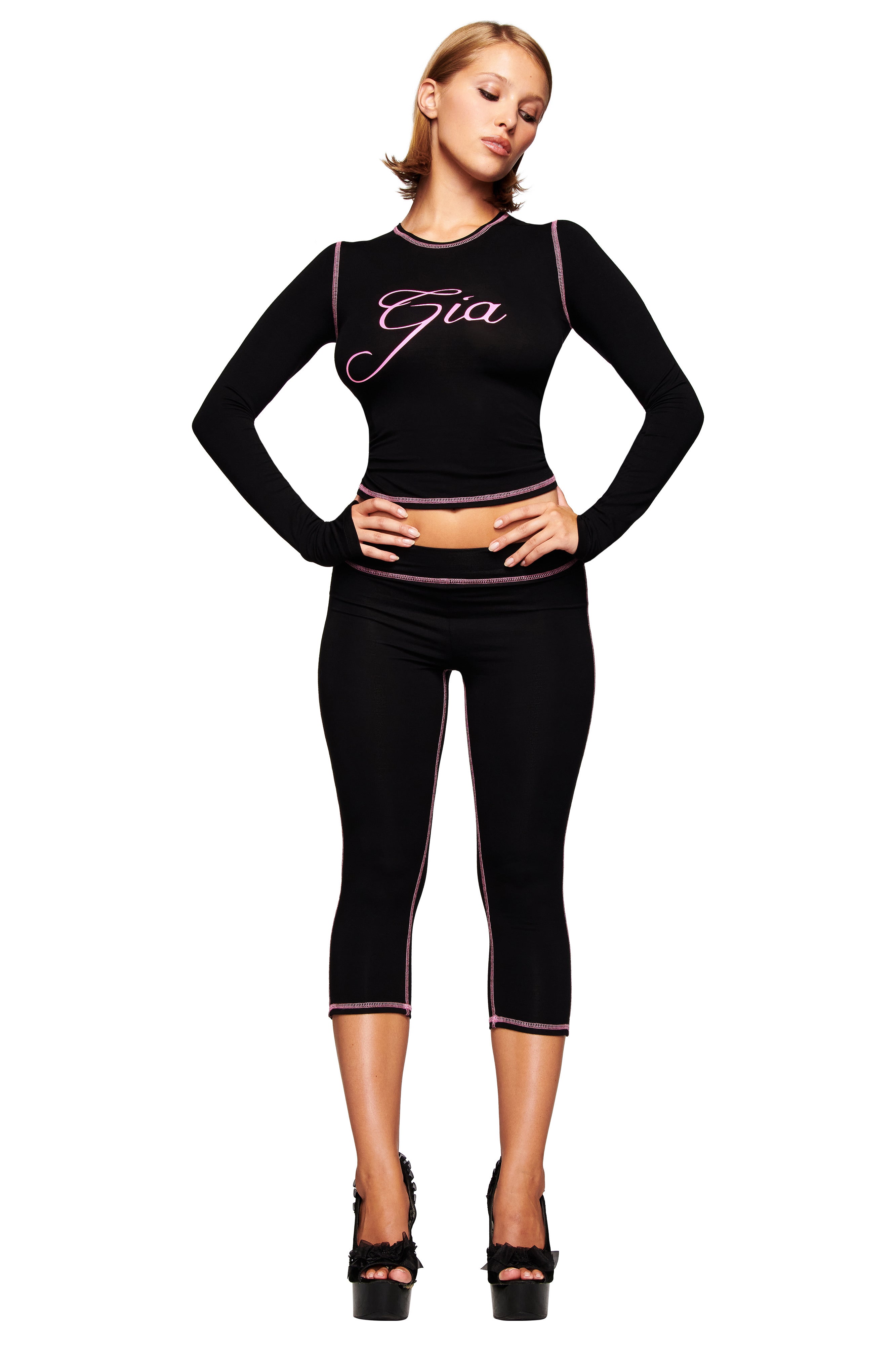 A woman stands with hands on her hips, wearing the I.AM.GIA BLARE LONG SLEEVE TOP - BLACK, matching black capri leggings, and black platform heels against a white background.