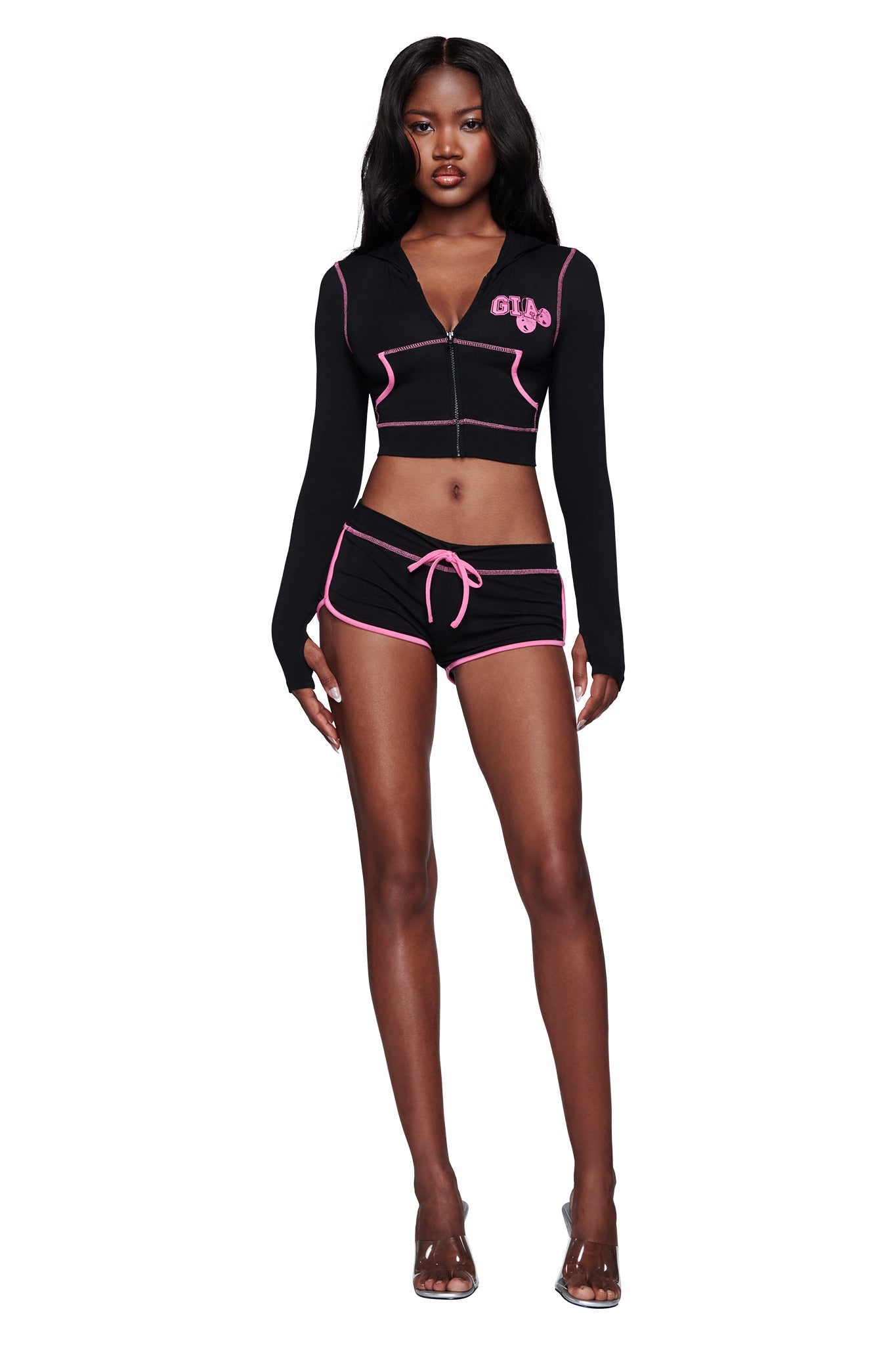 A woman stands against a white background wearing the I.AM.GIA BLARE CROPPED HOODIE in black, featuring pink trim and "GUU" text, paired with matching black shorts with pink trim and a tie, and clear high-heeled sandals.