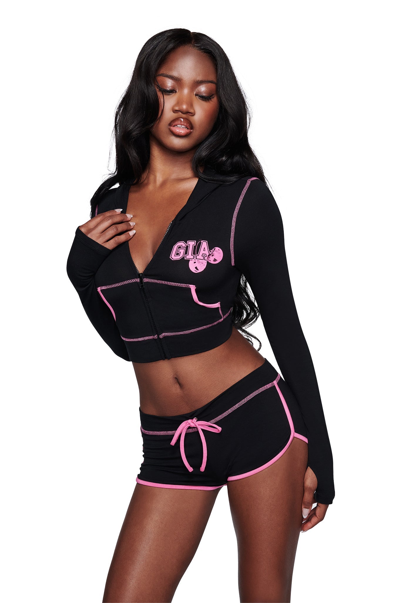 A woman poses in the I.AM.GIA BLARE CROPPED HOODIE in black, featuring pink trim and "GIA" printed on the left side, paired with matching black shorts with a pink drawstring. She stands against a white background.