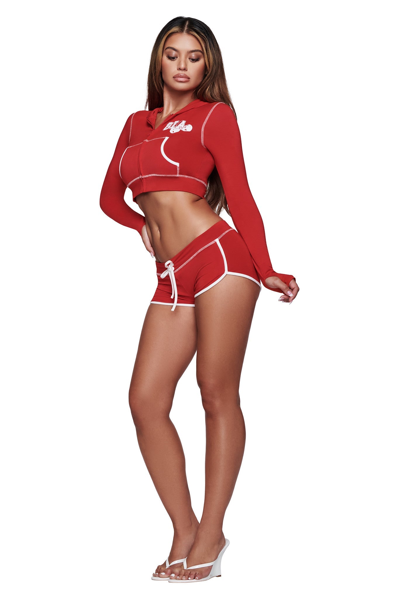 A woman poses against a white background wearing the I.AM.GIA BLARE CROPPED HOODIE in red, paired with matching red shorts and clear high-heeled shoes.