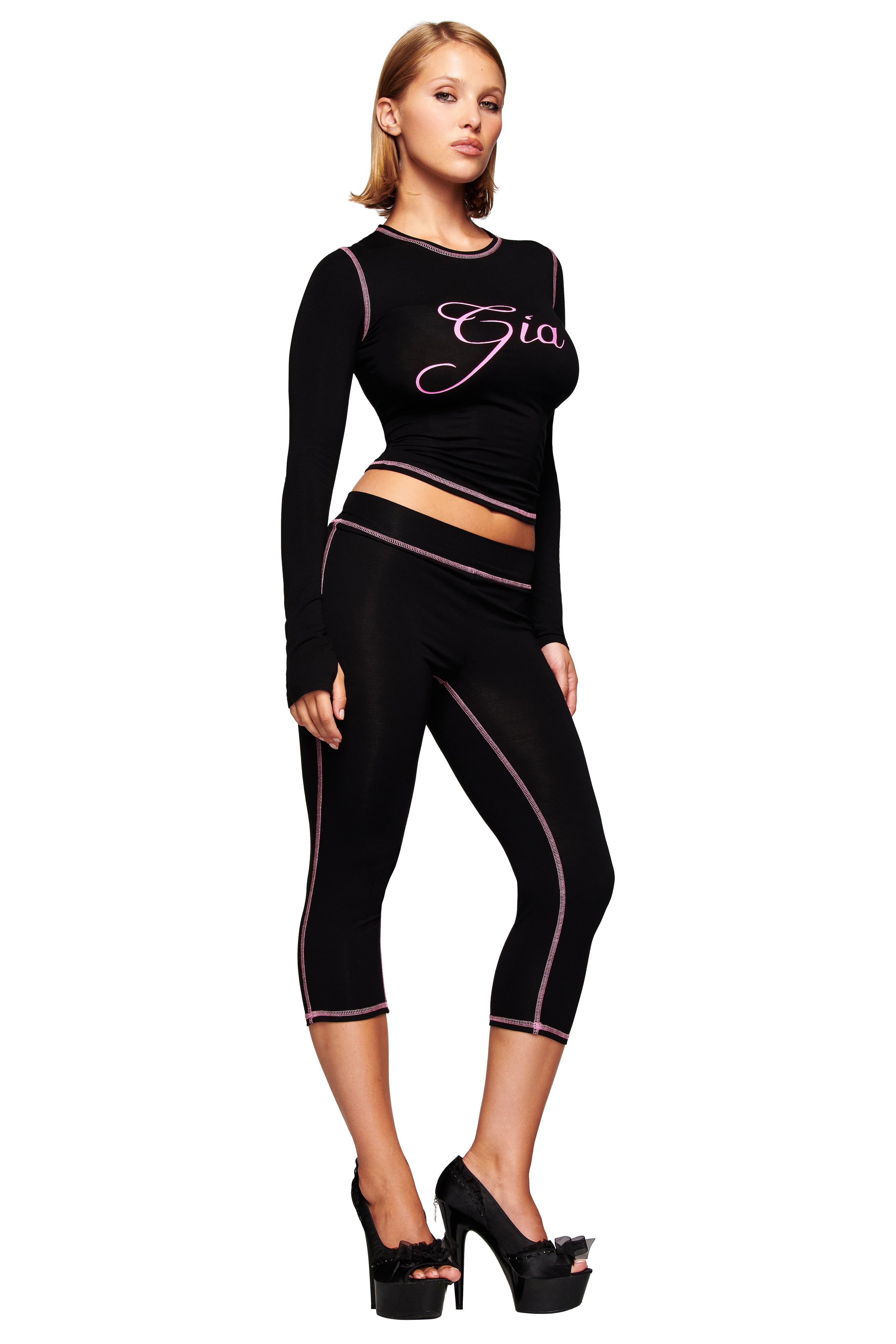 A woman poses against a white background wearing the black I.AM.GIA BLARE LONG SLEEVE TOP, paired with black capri leggings with pink trim and black platform high-heeled shoes.
