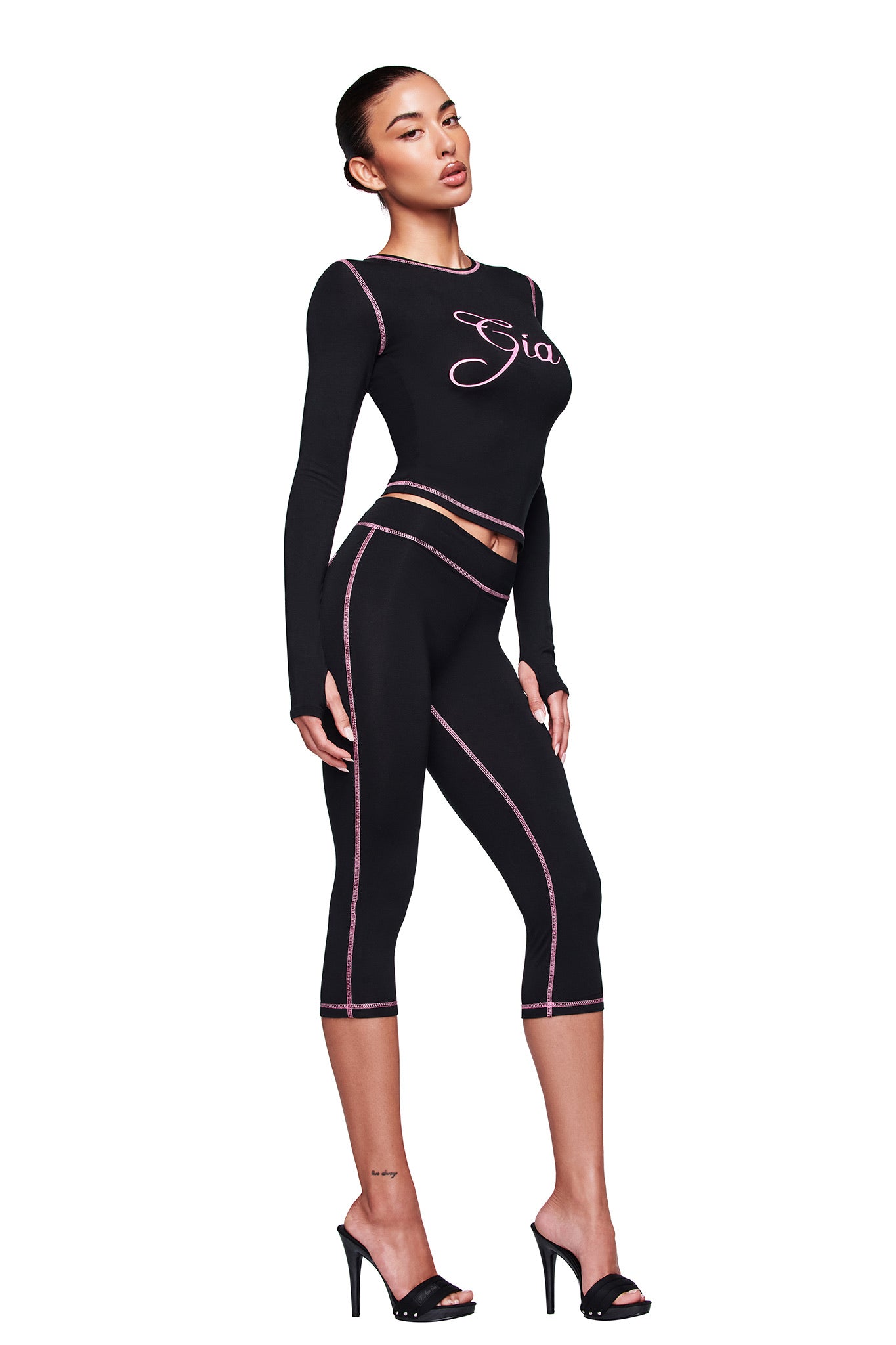 A woman models the I.AM.GIA BLARE CAPRI - BLACK leggings with pink stitching and a matching fitted black I.AM.GIA crop top, paired with black heels, all set against a white background.