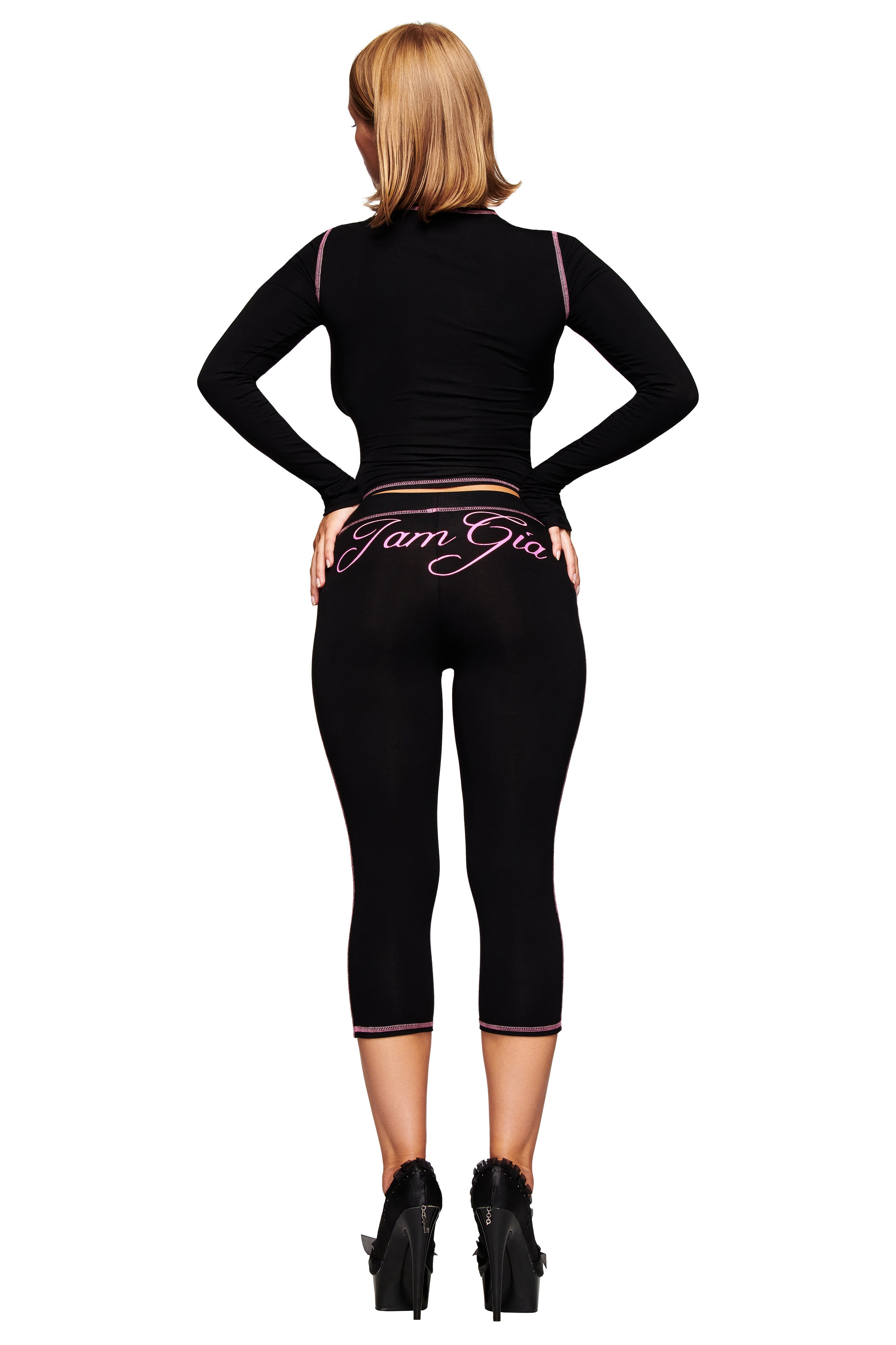 A woman faces away wearing a black long-sleeve top and I.AM.GIA BLARE CAPRI - BLACK pants featuring the brand's pink script logo on the back, paired with black high heels.
