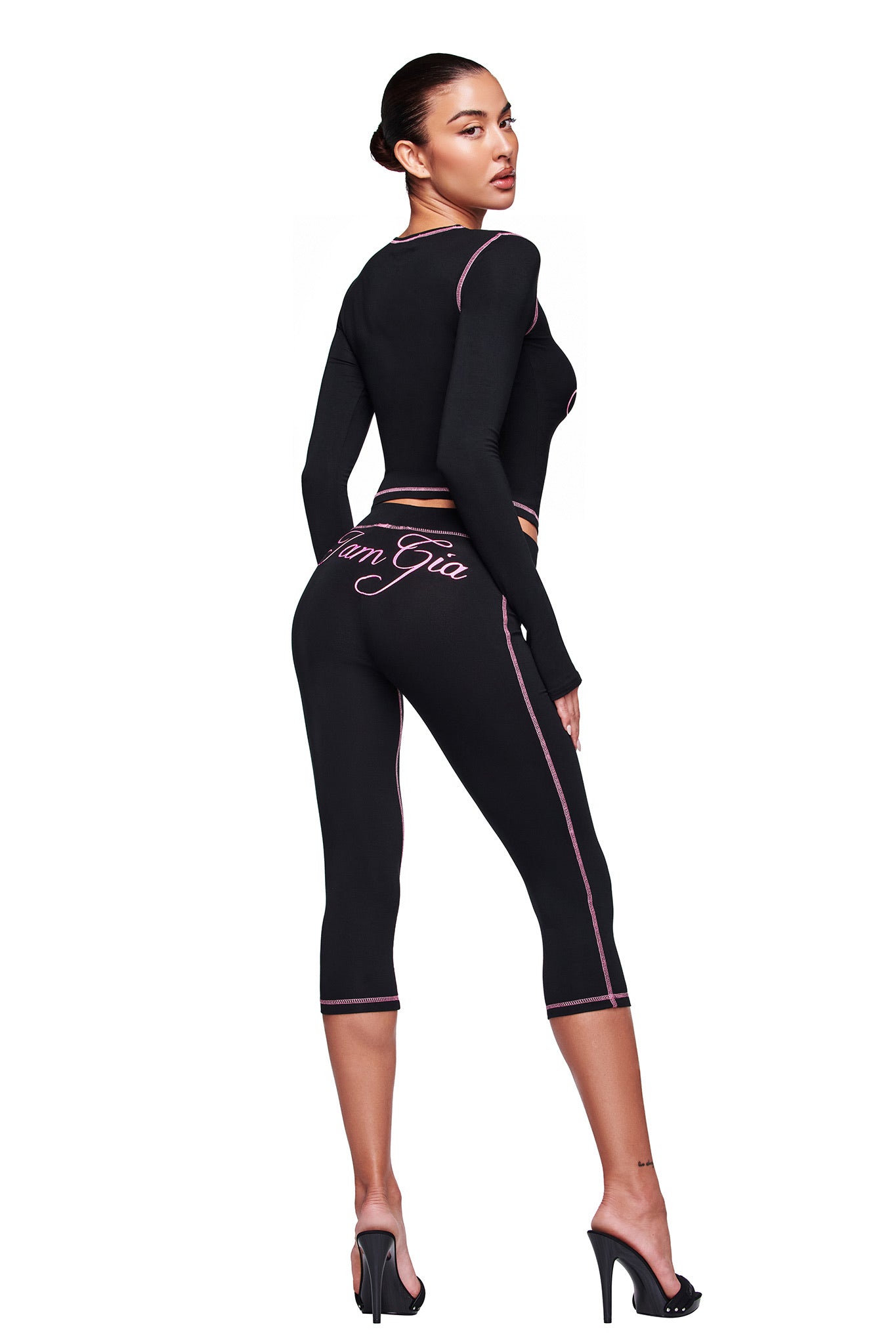 A woman wears the I.AM.GIA BLARE CAPRI - BLACK, featuring pink trim and writing on the waistband, as she stands in high heels and glances over her shoulder against a white background.