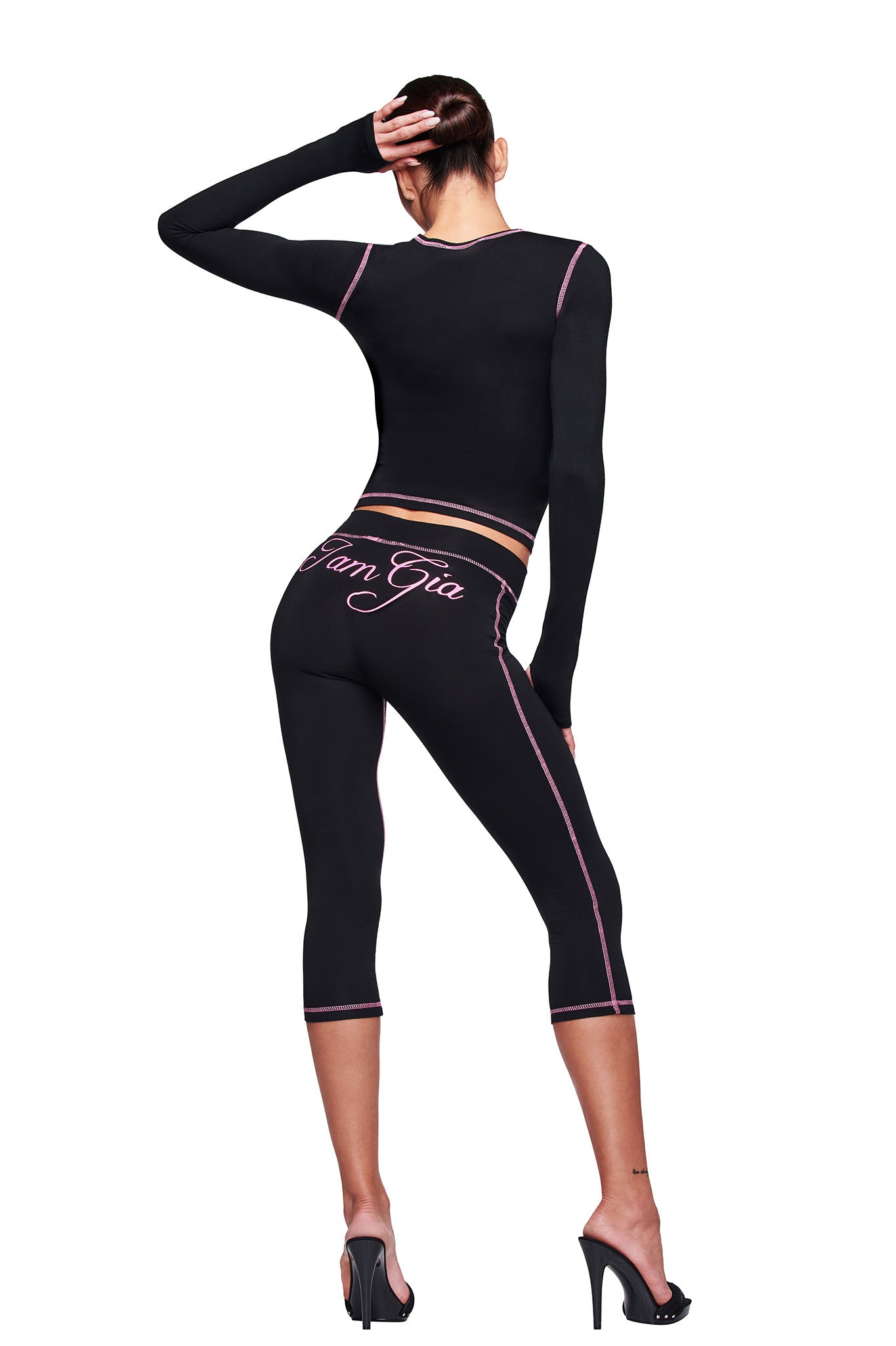 A woman wears the I.AM.GIA BLARE LONG SLEEVE TOP in black with thumb holes and matching cropped leggings featuring pink seams and "Tam Gia" on the back, paired with black high-heeled sandals.
