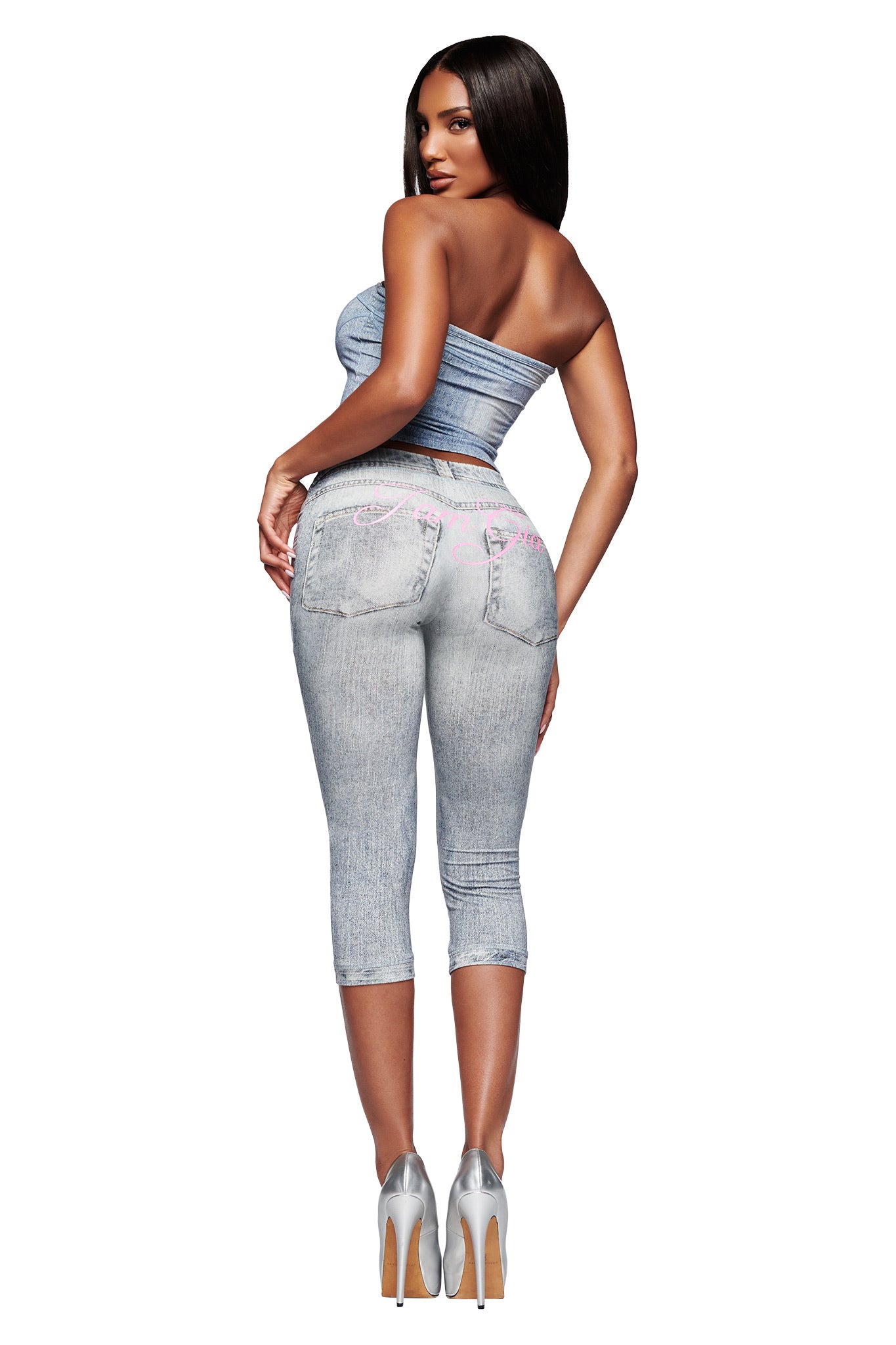 A woman with long dark hair wears the I.AM.GIA BLARE BOOB TUBE - DENIM TROMPE L'OEIL, light-wash embroidered capri jeans, and silver heels. She stands facing backward, looking over her shoulder.
