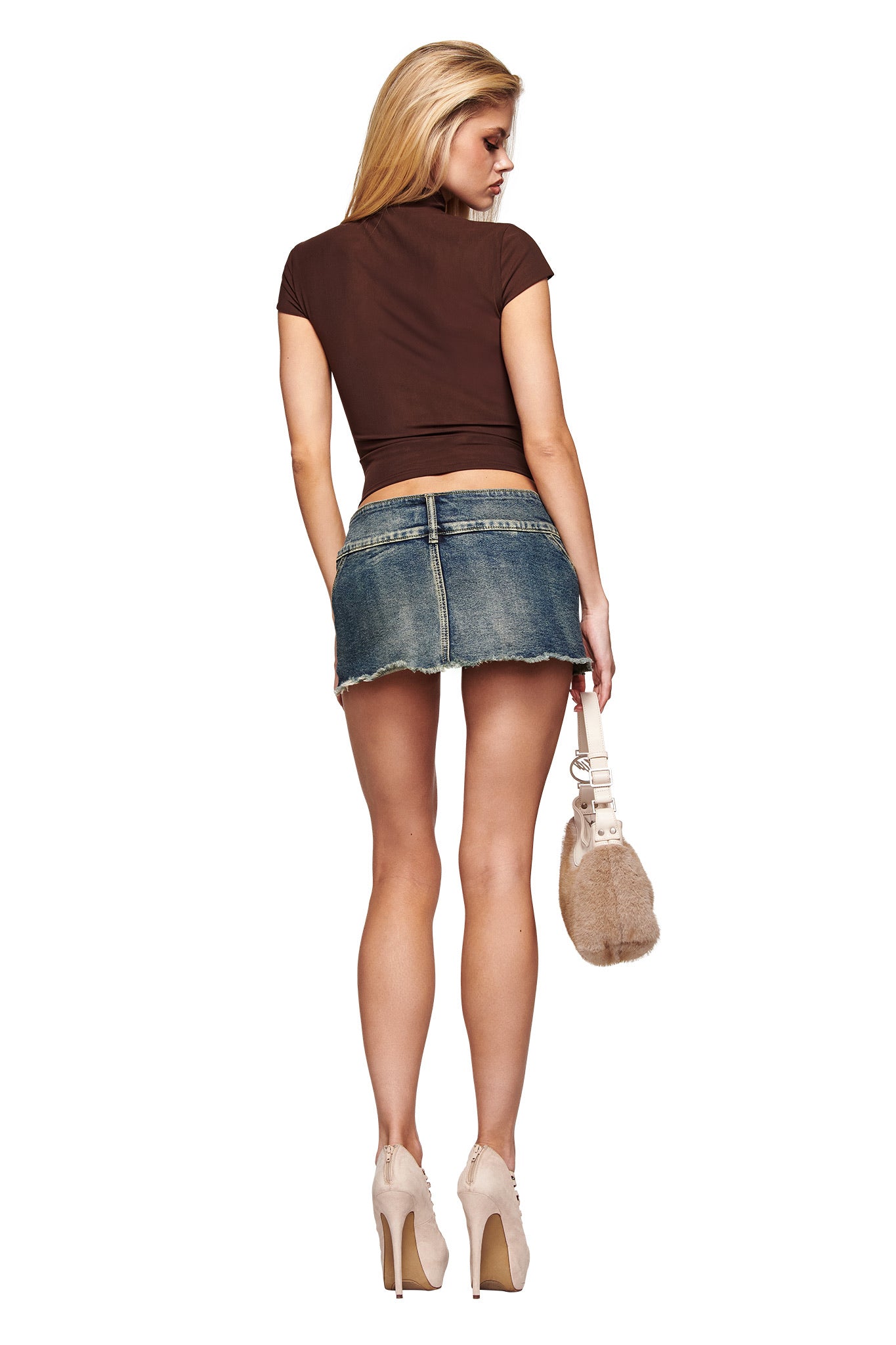 A woman with long blonde hair faces away, wearing the I.AM.GIA ISELA TOP in brown with short sleeves, a bodycon denim mini skirt, beige high heels, and a small beige handbag against a plain white background.