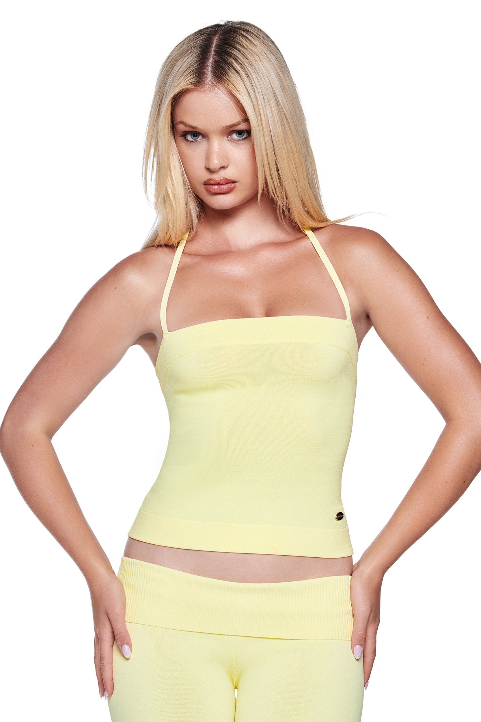 A woman with long blonde hair stands against a white background, wearing the I.AM.GIA AUDRINA TOP in baby yellow and matching high-waisted pants, hands on her hips.