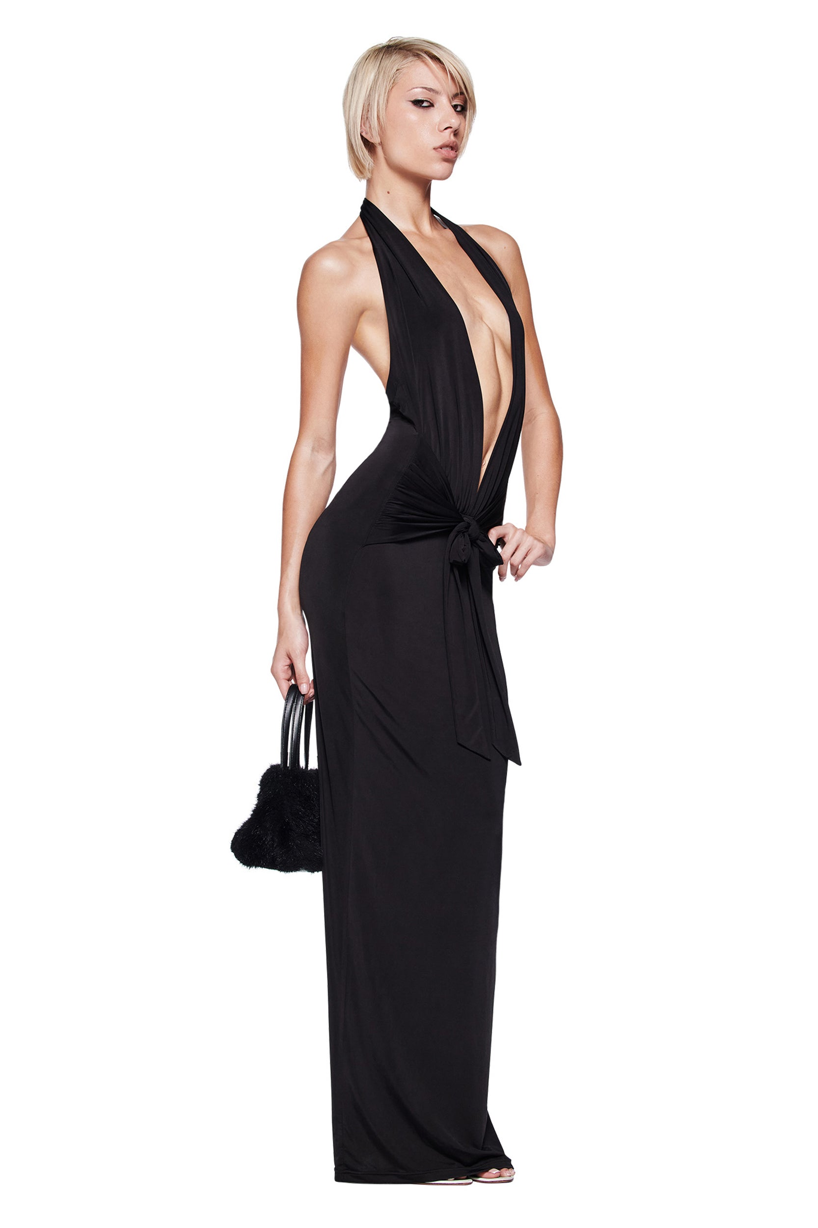 A woman with short blonde hair models the I.AM.GIA ZIVA PLUNGE TIE MAXI DRESS in black, featuring a bodycon fit and plunging V neckline. She holds a small black handbag and stands confidently against a white background.