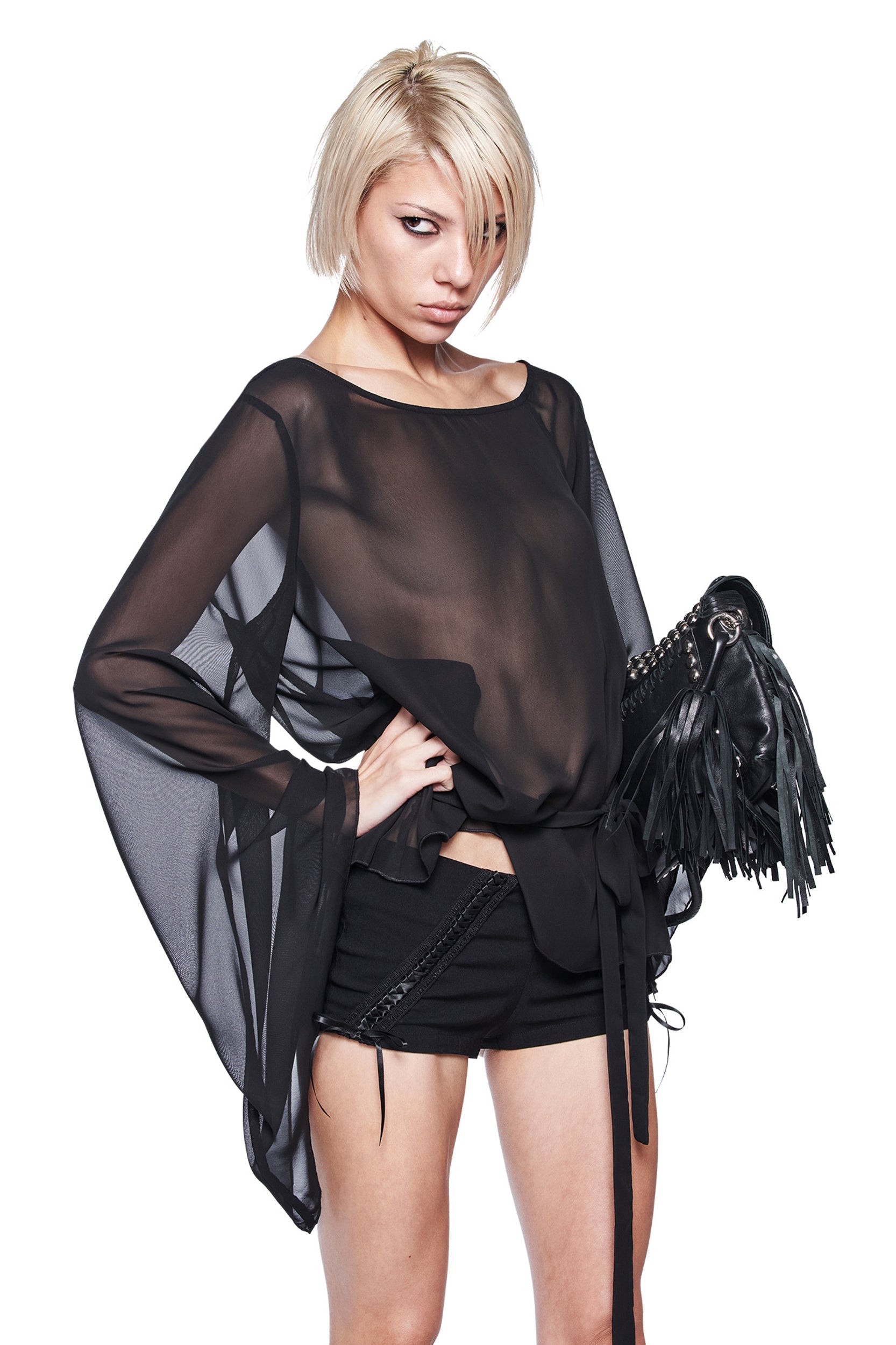 A person with short blonde hair wears the I.AM.GIA MALINA LONG SLEEVE PONCHO TOP and black shorts, posing with one hand on their hip and holding a fringed black handbag against a plain white background.