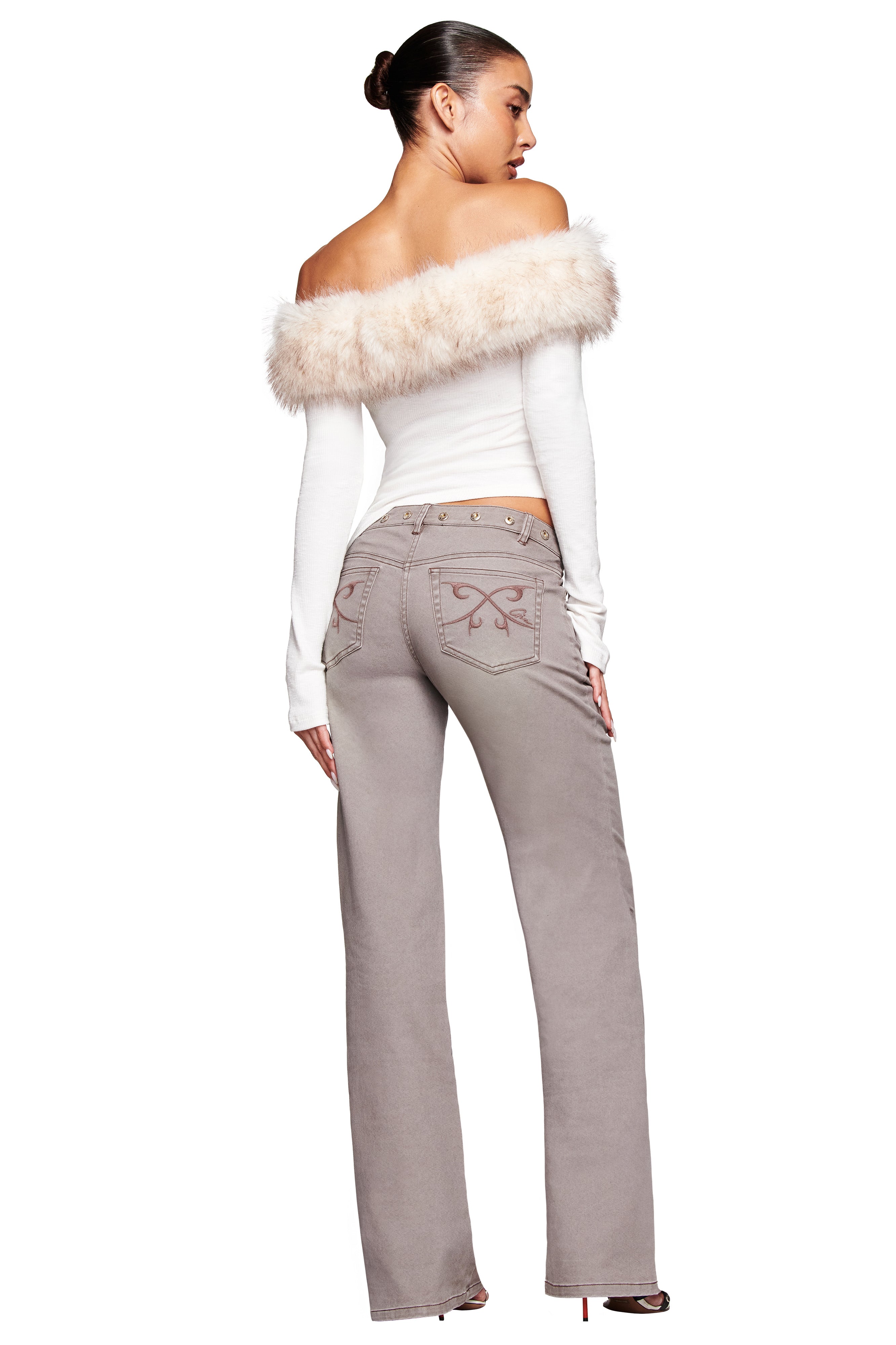 A woman faces away wearing the I.AM.GIA CECILY TOP in cream, featuring a ribbed off-shoulder design with a removable faux fur collar, styled with high-waisted taupe jeans and heeled shoes. Her hair is in a sleek bun against a white background.