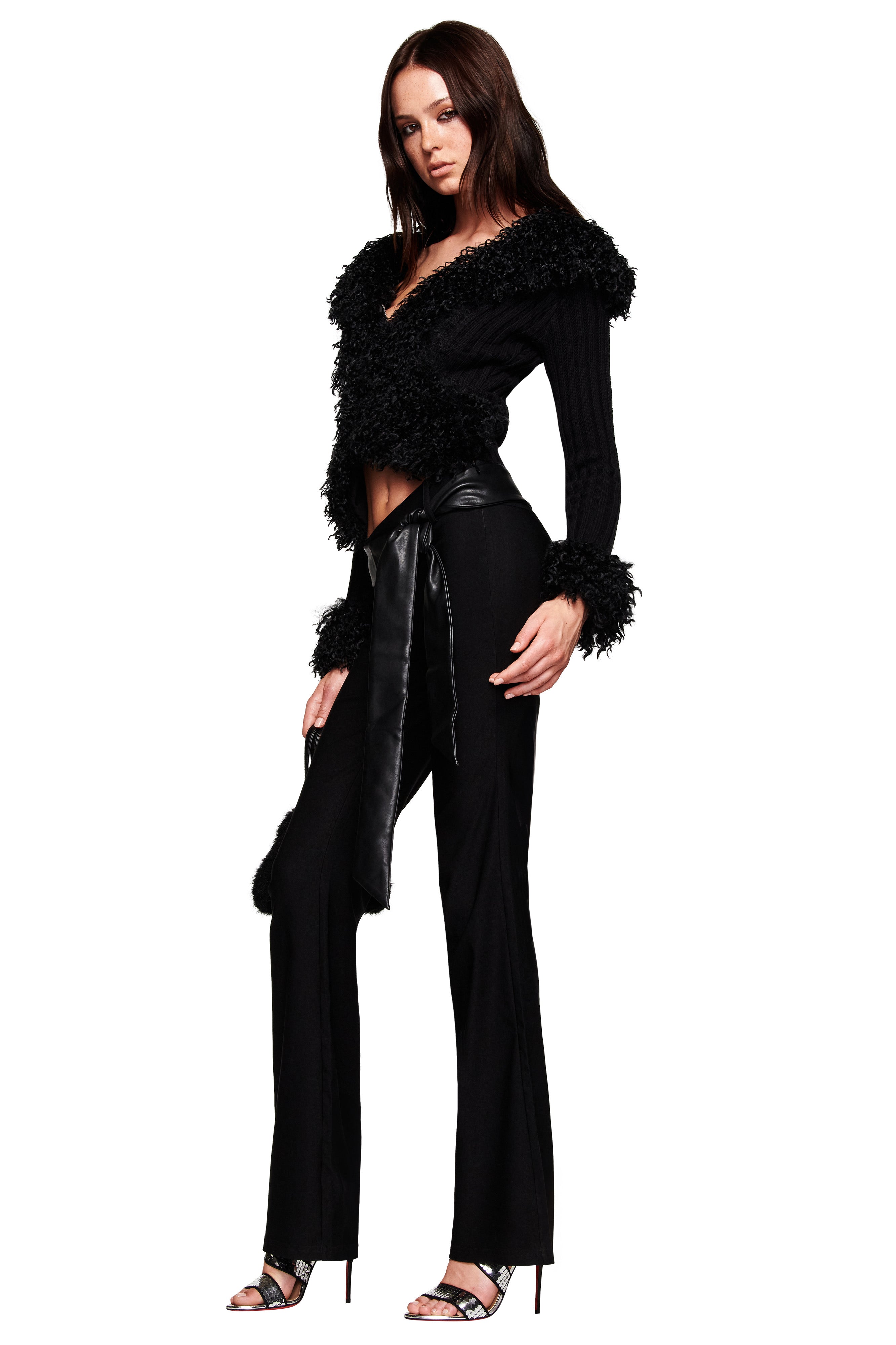A woman poses confidently in I.AM.GIA's FELIZ PANT - BLACK, styled with a furry wrap top, faux leather belt, and high heels against a white background.
