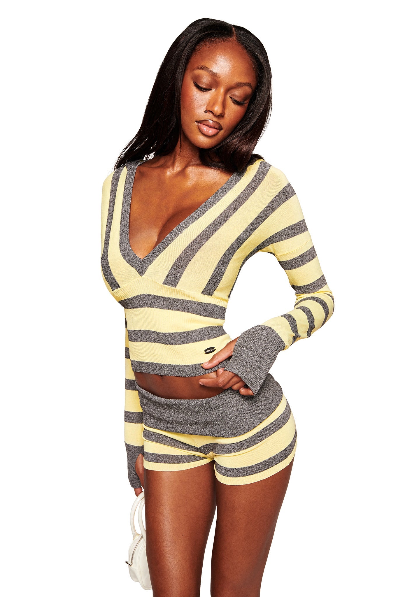 A woman wears the I.AM.GIA SUZI SWEATER in yellow and grey stripe with matching shorts, posing with one hand on her hip, eyes closed. She holds a small white bag and has long straight hair against a white background.