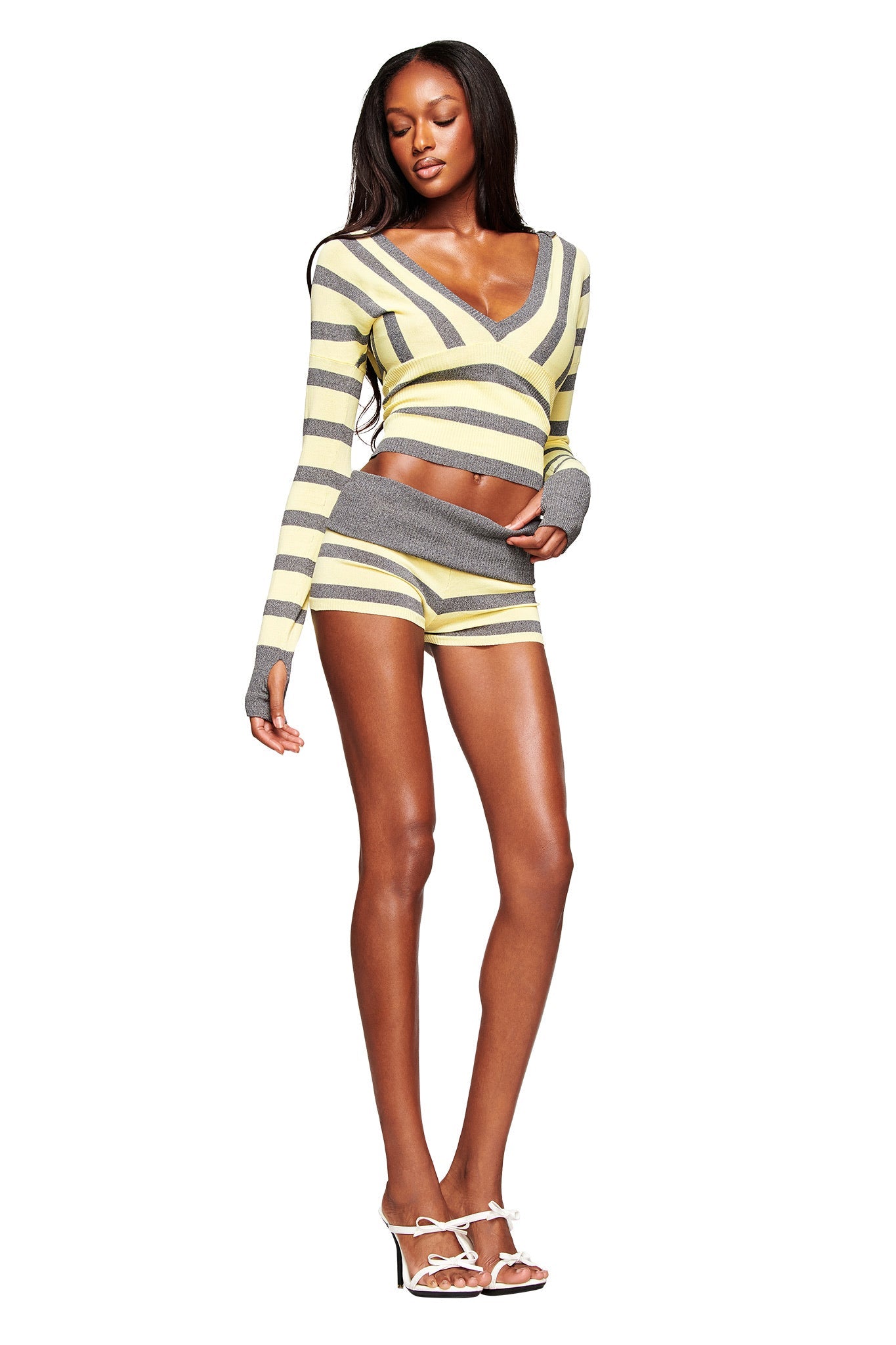 A woman poses against a white background wearing the I.AM.GIA SUZI SHORT - YELLOW AND GREY STRIPE in a micro mini length, paired with a matching striped long-sleeve crop top and white high-heeled sandals.