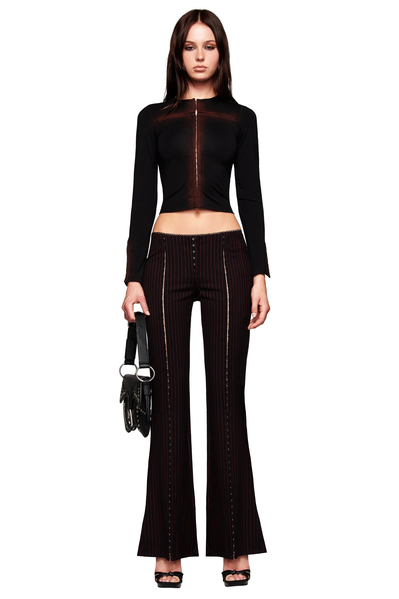 A woman faces forward in a long-sleeve black crop top and I.AM.GIA RONAN PANT in black/red pinstripe, holding a black handbag and wearing black open-toe heels against a white background.