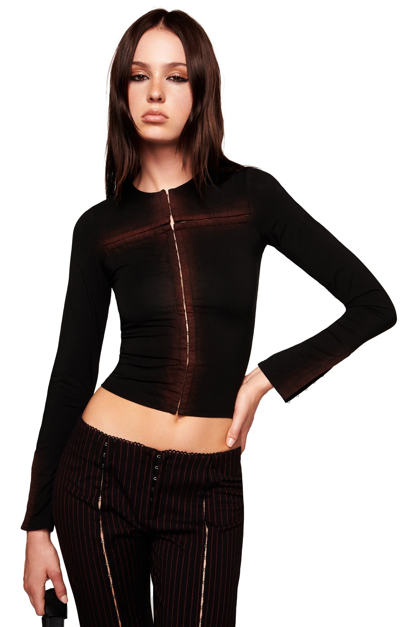 A woman with long dark hair poses against a white background, wearing I.AM.GIA's RONAN PANT in black/red pinstripe with a straight leg, paired with a fitted black zip-front long-sleeve top and one hand on her hip.