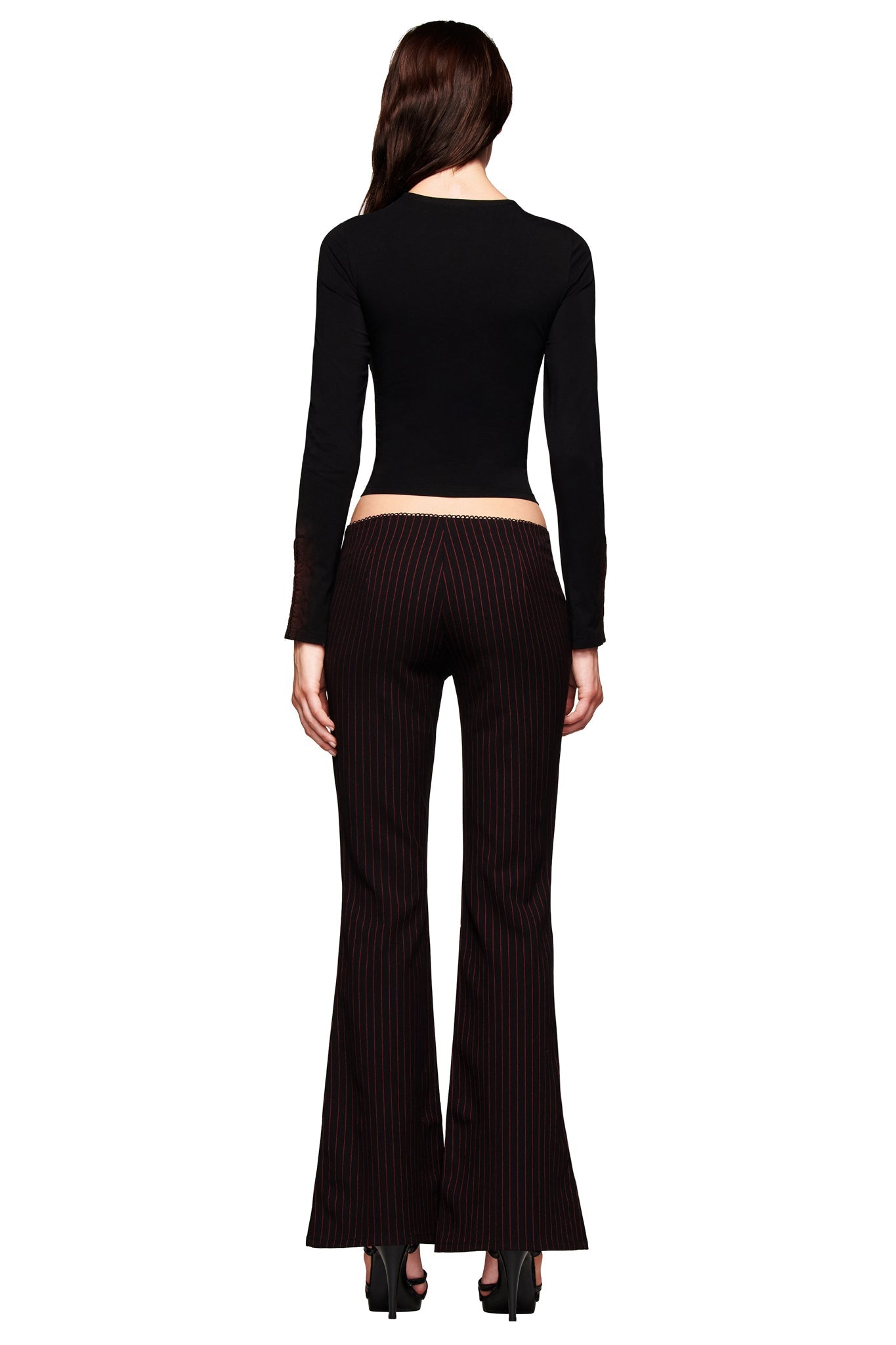 A woman faces away, wearing a fitted black long-sleeve top and the I.AM.GIA RONAN PANT in black/red pinstripe with black high-heeled shoes, set against a white background.