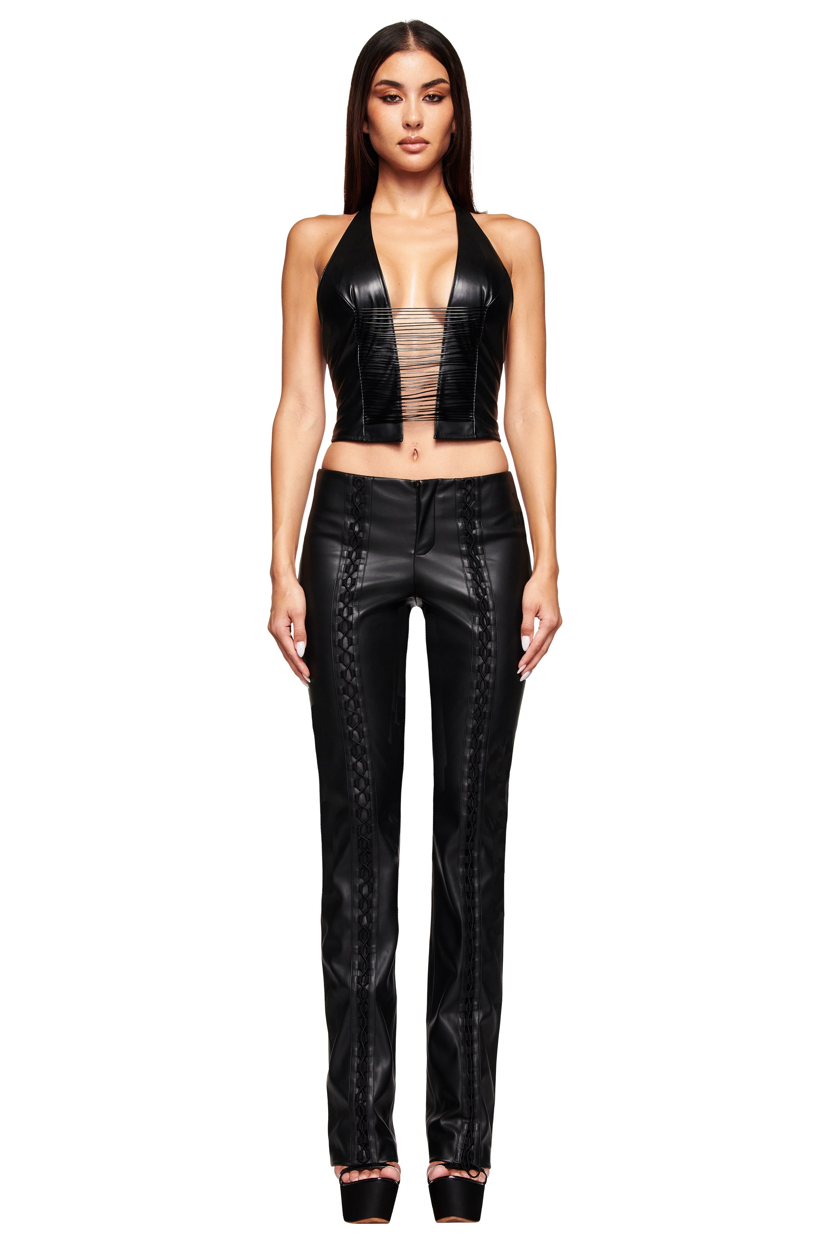 A woman with long dark hair and neutral makeup poses against a white background, wearing the I.AM.GIA ALTAIR TOP - BLACK, styled with black bodycon leather pants, a matching sleeveless vest, and high platform heels.