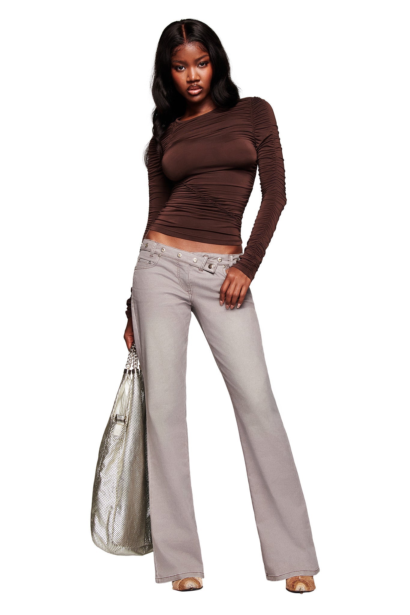 A woman stands confidently in I.AM.GIA's GREER PANT - GREY, featuring a V-shaped waistband and flared legs. She pairs them with a fitted brown long-sleeve top, holds a large metallic silver bag, and has long wavy hair.