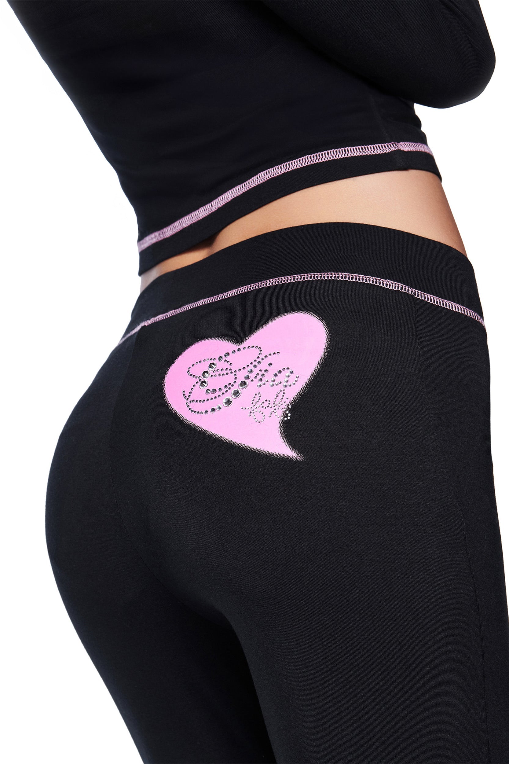 A person wears the I.AM.GIA FAFI BLARE PANT in black, featuring a pink heart with rhinestones on the back, paired with a matching black top with pink trim. The photo is cropped to show only the lower back and upper leg.