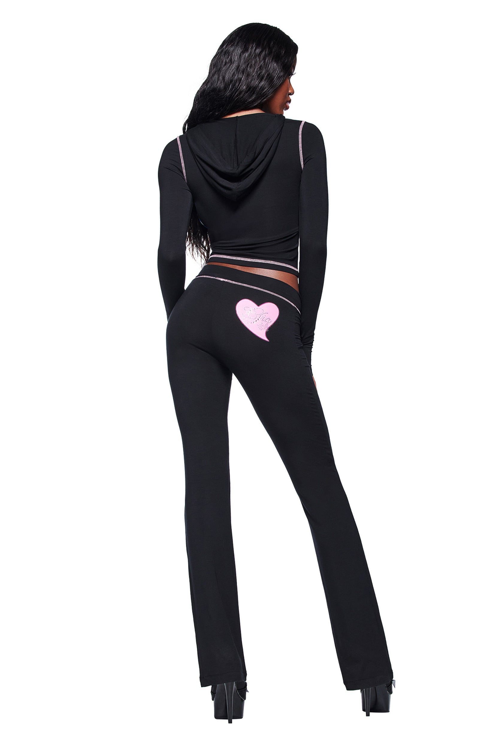 A woman with long dark hair wears the I.AM.GIA FAFI BLARE HOODIE in black, featuring pink accents, diamanté details, and a pink heart on the back of the pants. She faces away and pairs the tracksuit with high-heeled shoes.