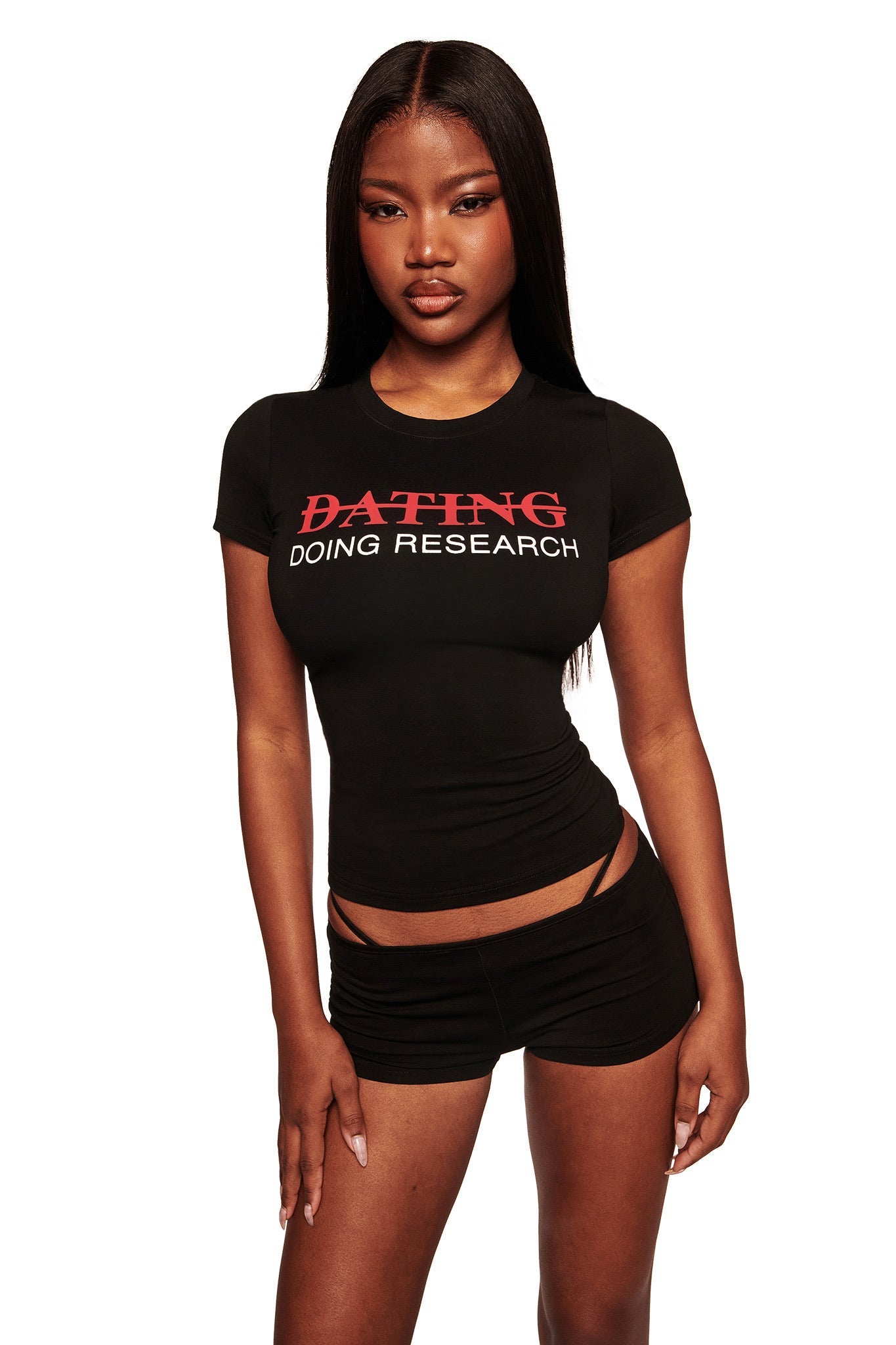 A woman with long straight hair faces the camera in a fitted black I.AM.GIA POLINA TEE - RESEARCH graphic t-shirt and black cut-out strap shorts, set against a white background.