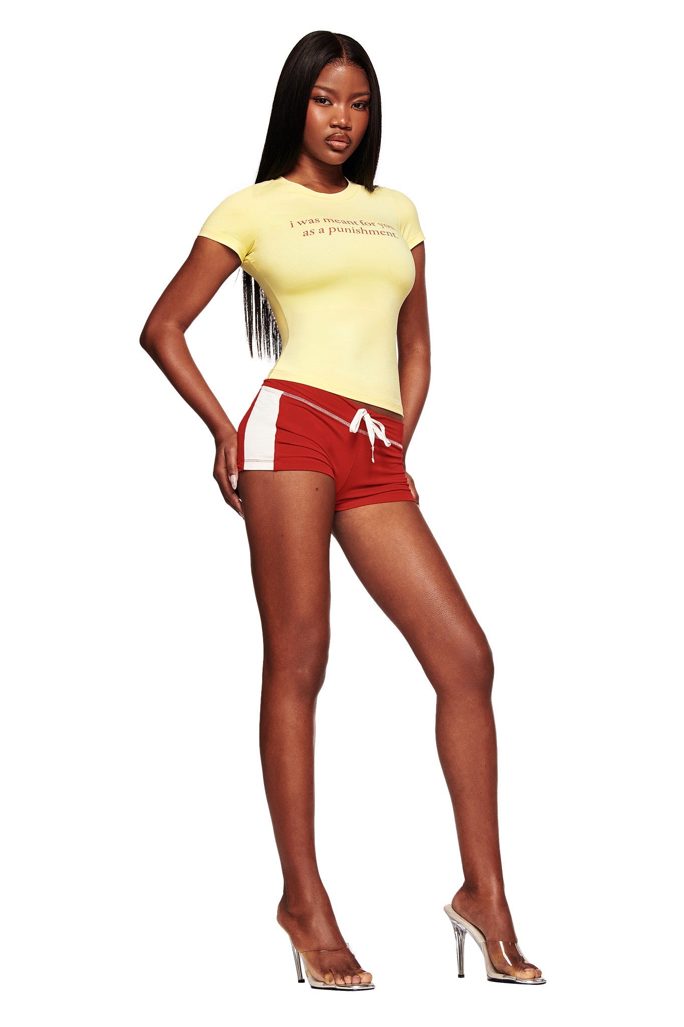 A woman with long straight hair and a neutral expression wears the I.AM.GIA POLINA TEE - PUNISHMENT in yellow, paired with red athletic shorts featuring white side panels and clear high-heeled shoes.