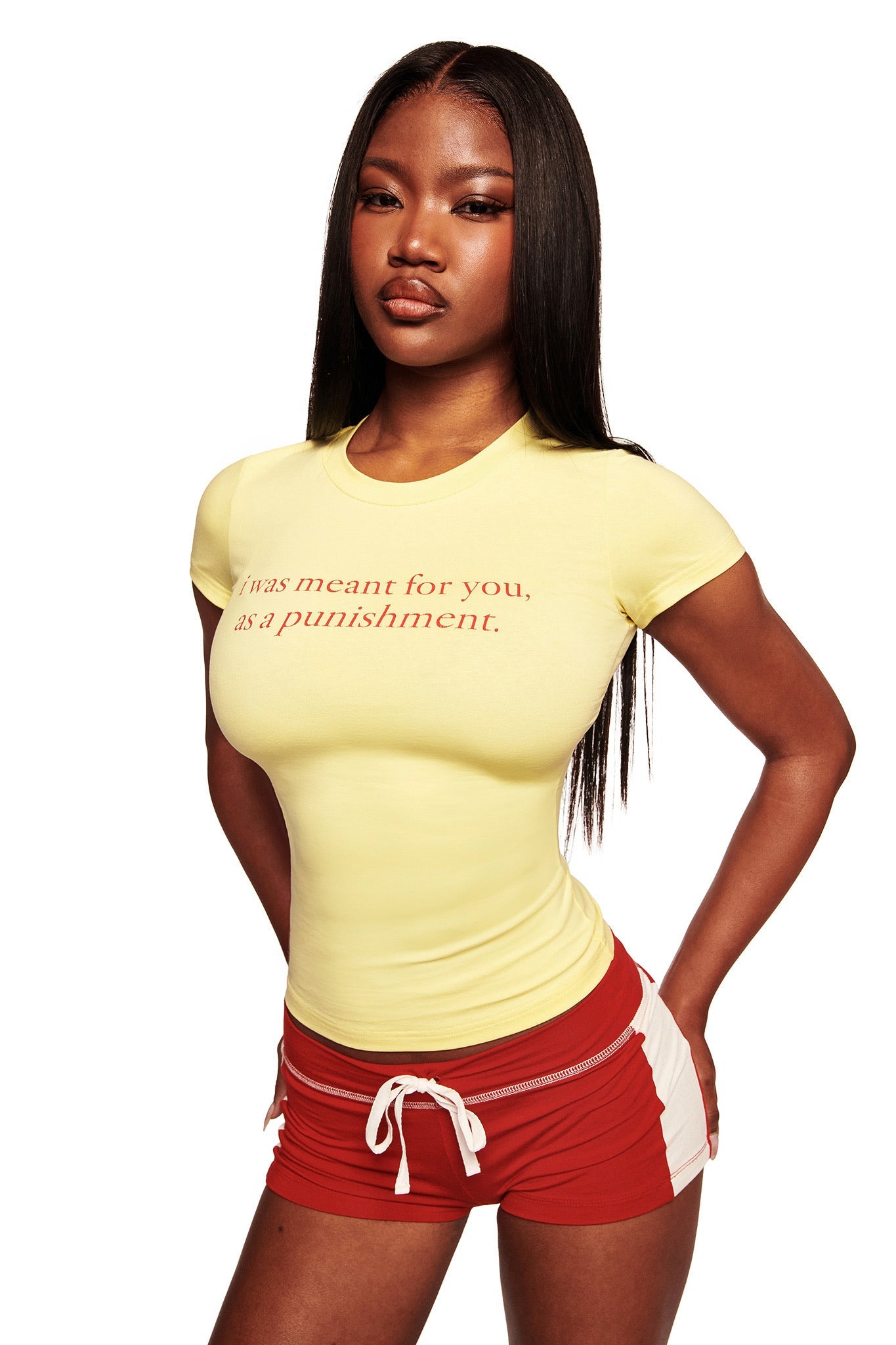A woman with long straight hair wears the I.AM.GIA POLINA TEE - PUNISHMENT, a fitted yellow graphic t-shirt reading "I was meant for you, as a punishment," paired with red athletic shorts, standing against a white background.