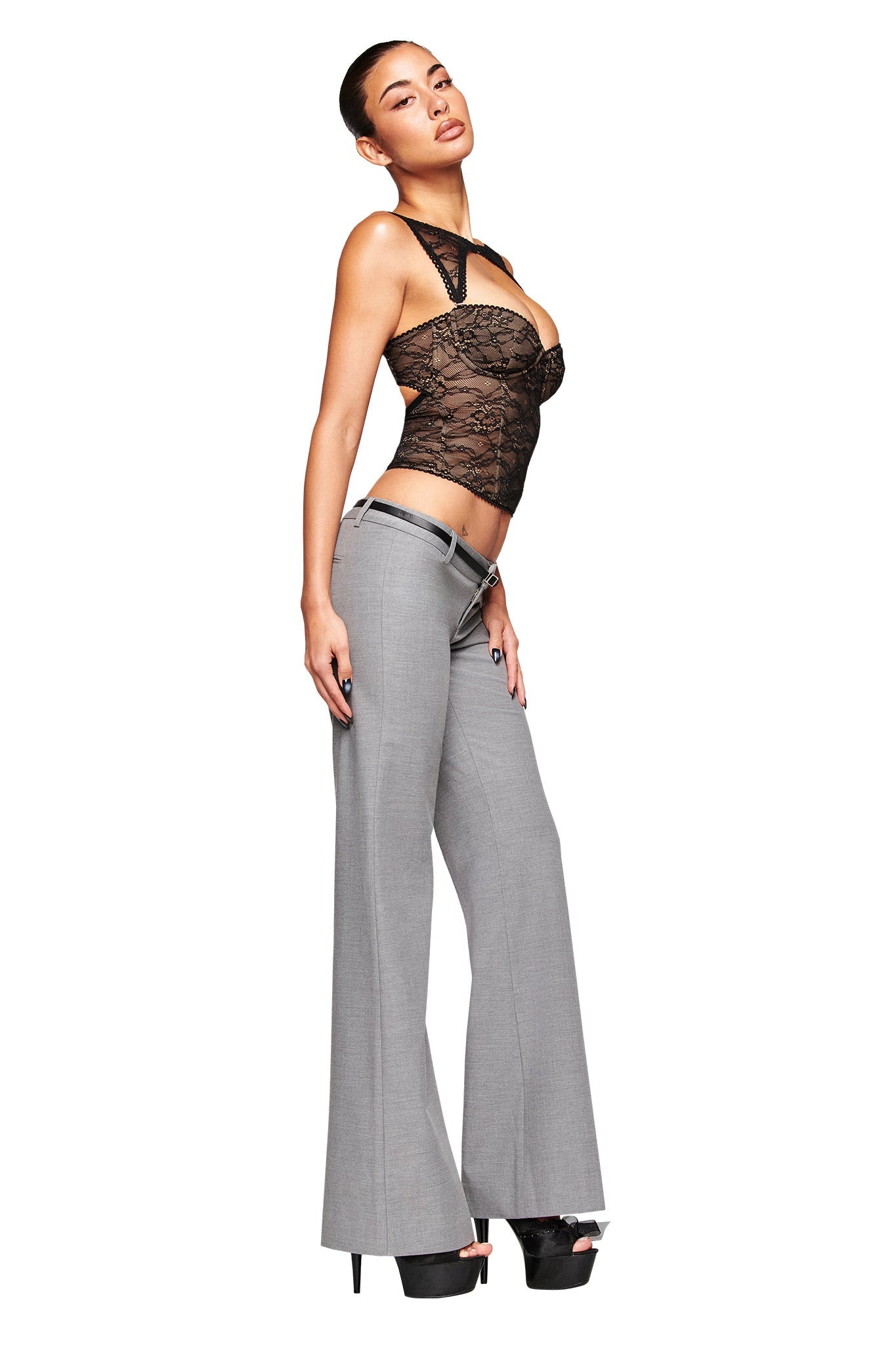 A woman poses confidently in a sheer black lace top, I.AM.GIA’s CLARA PANT in grey with a relaxed straight leg, and black high platform heels against a white background.