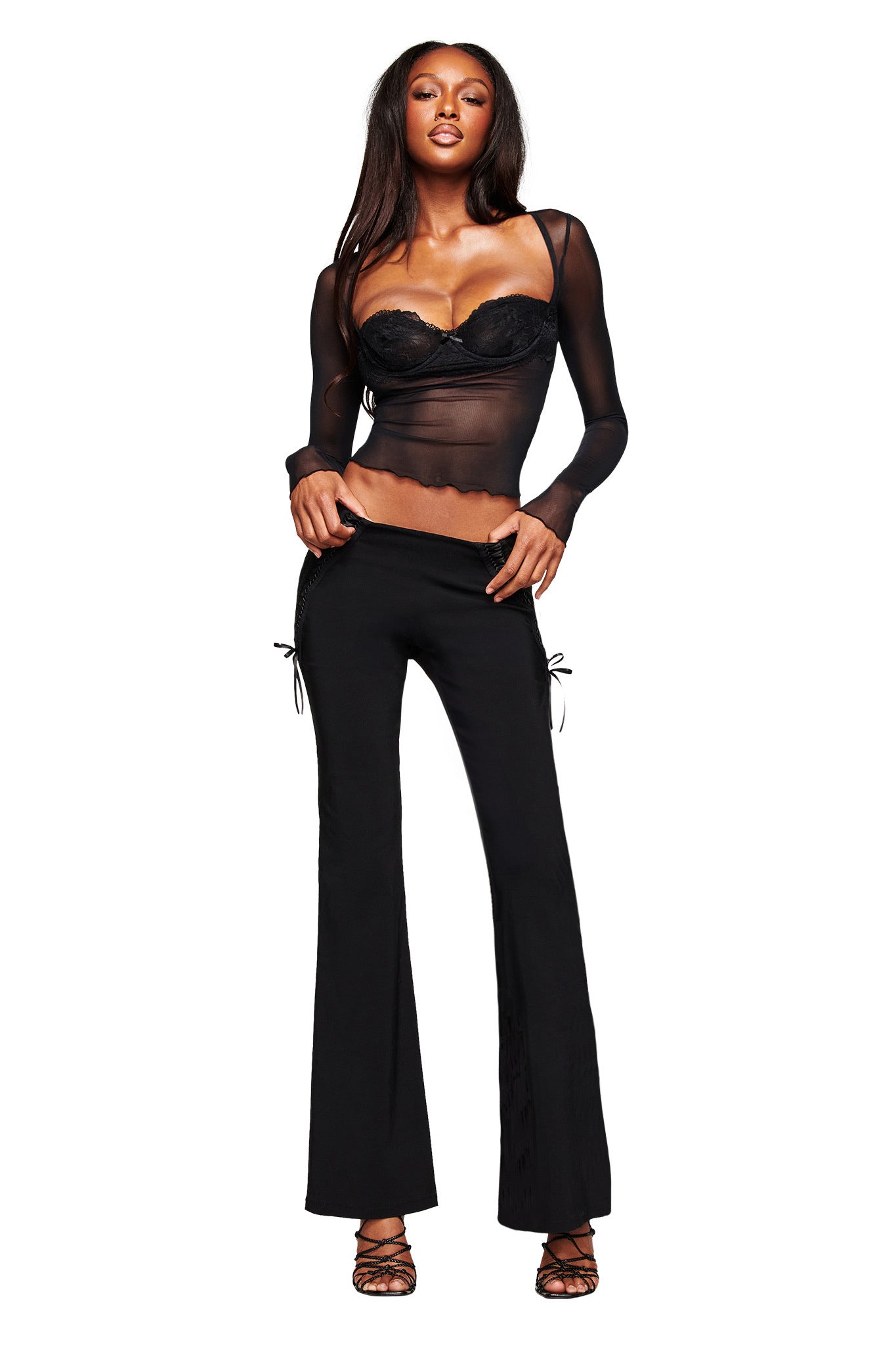 A woman poses confidently in the I.AM.GIA SILO LONG SLEEVE TOP - BLACK, layered over a black lace bustier, paired with black flared pants featuring side ties and strappy heels. Her long hair and bold stance add to her striking look.