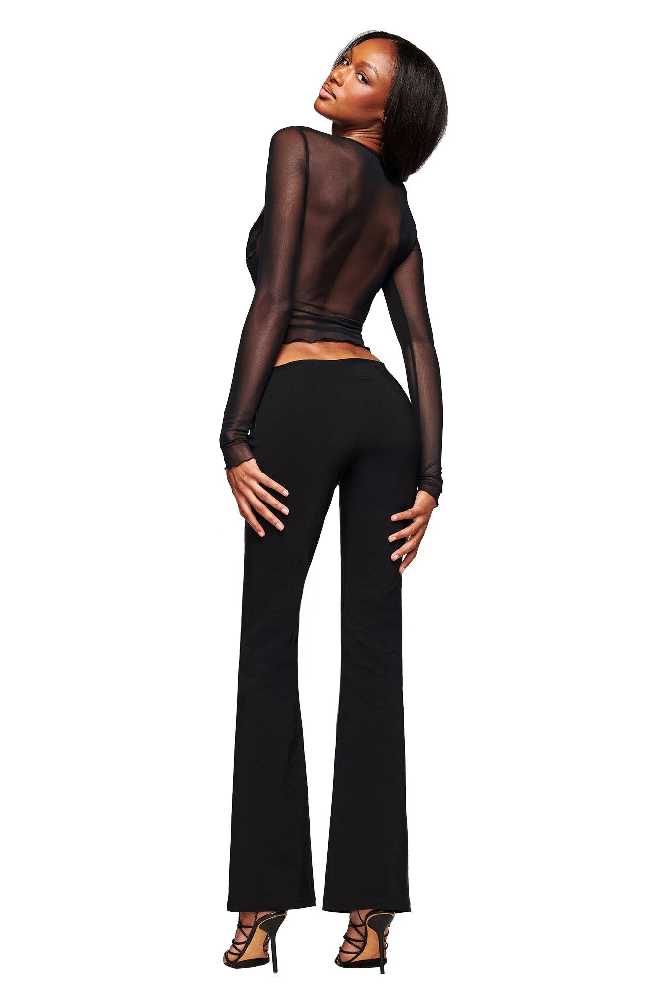 A woman poses with her back slightly turned, wearing the I.AM.GIA SILO LONG SLEEVE TOP in black, paired with black pants and high-heeled sandals against a plain white background.