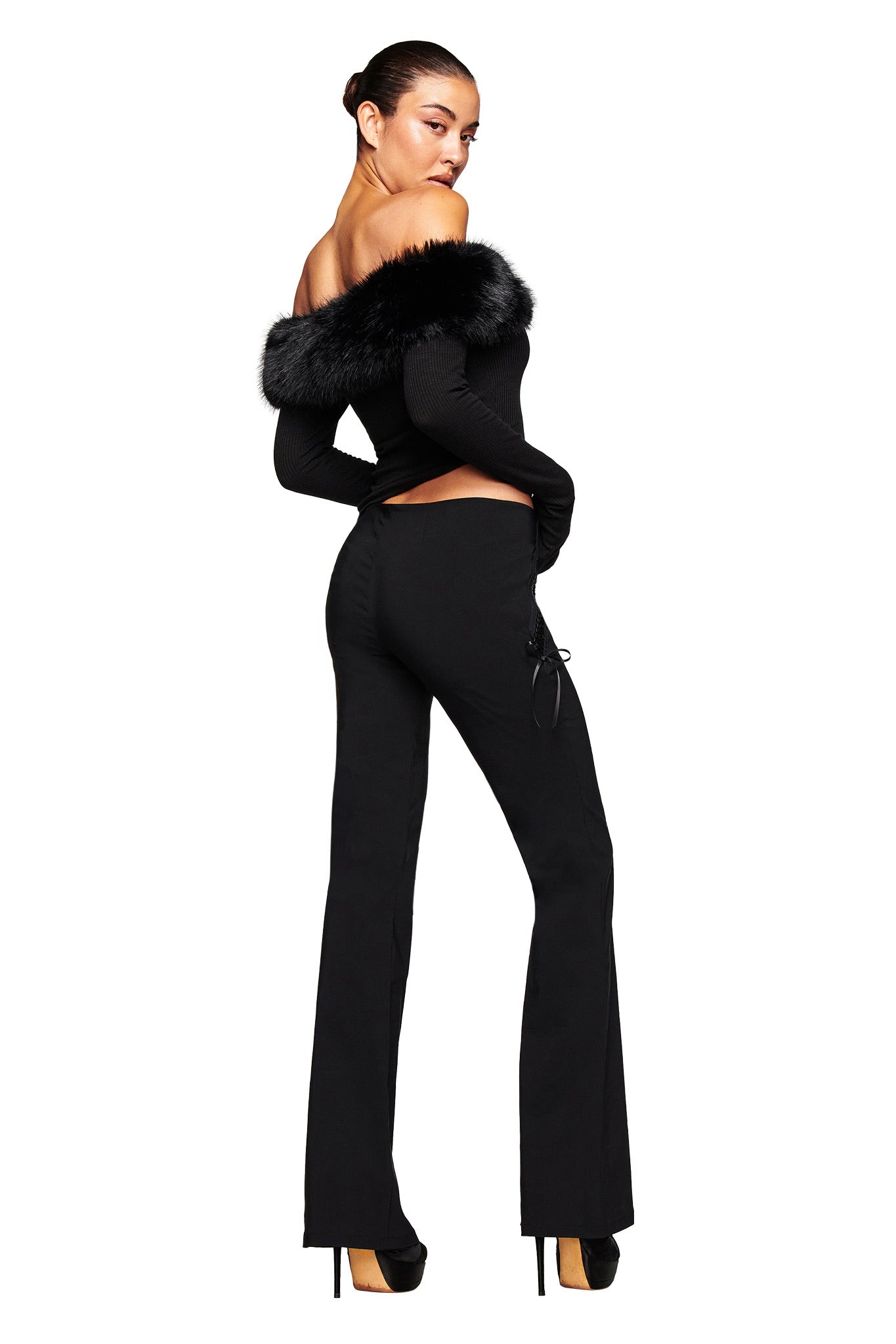 A woman models the I.AM.GIA SAMIRA PANT in black, paired with a black off-the-shoulder faux fur trim top and high heels. She stands against a white background, turned away and glancing over her shoulder.
