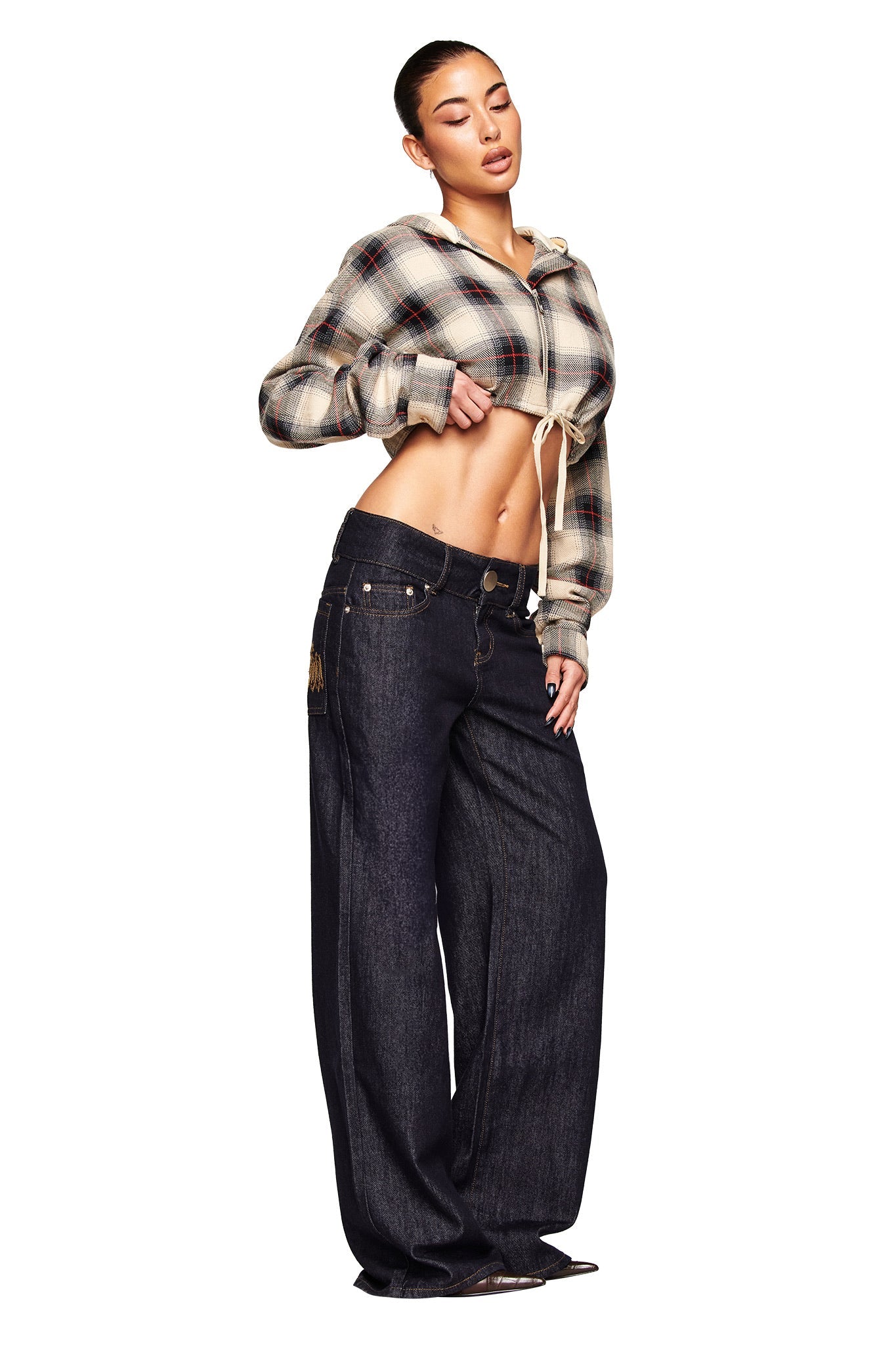 A woman poses against a white background wearing I.AM.GIA’s MOXIE JEAN - INDIGO, styled with a cropped plaid jacket. She stands confidently, showcasing the low-rise, loose-fit denim and revealing her midriff.