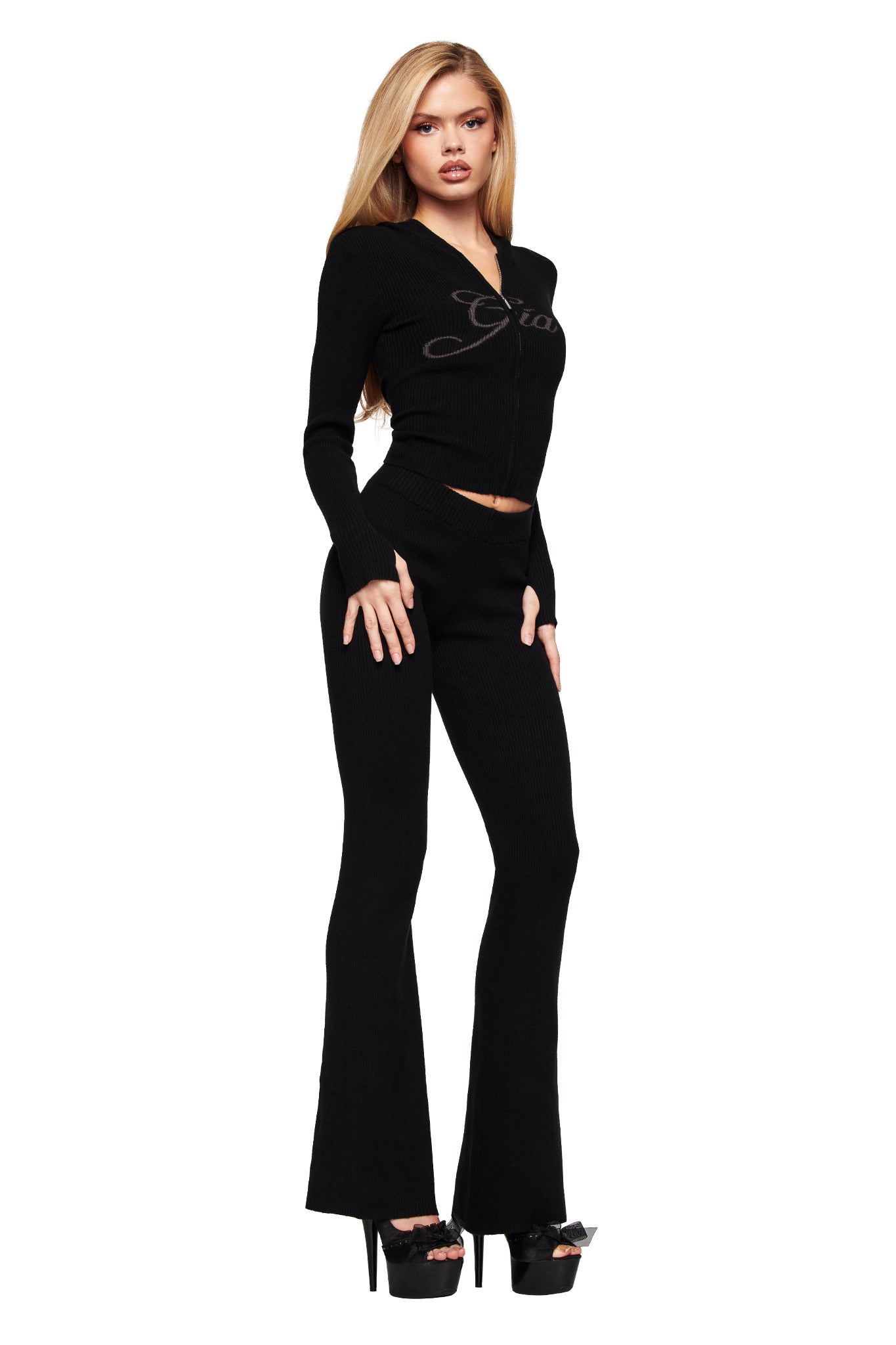 A woman with long blonde hair stands confidently against a white background wearing the I.AM.GIA BLARE KNIT TRACKPANT in black, featuring a mid-rise elasticated waistband and flared legs, paired with a fitted zip-up top and platform sandals.