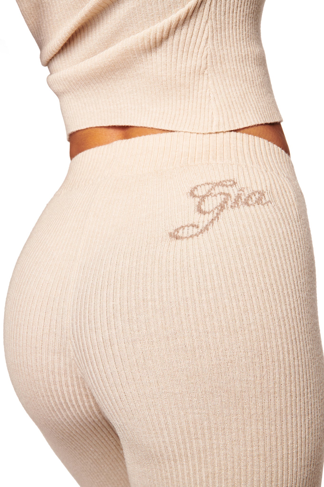 A close-up of a person wearing I.AM.GIA's BLARE KNIT TRACKPANT in cream, featuring a mid-rise elasticated waistband, gentle flare leg opening, textured fabric, and “Gia” embroidered near the waistband.