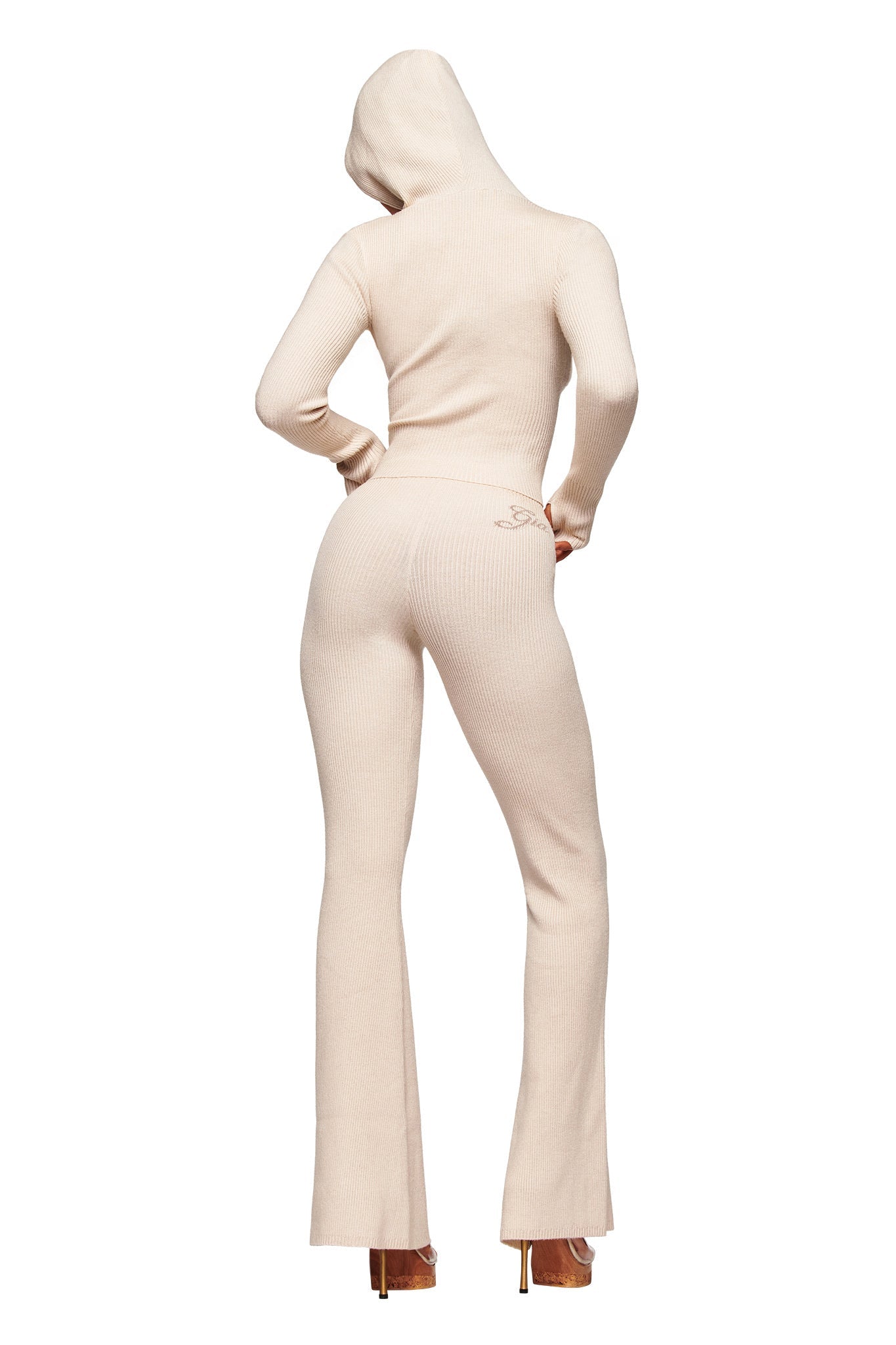 A person wearing the I.AM.GIA BLARE KNIT TRACKPANT in cream, featuring a fitted ribbed design, elasticated mid rise waistband, and flared legs, stands with their back to the camera and hands on hips against a white background.