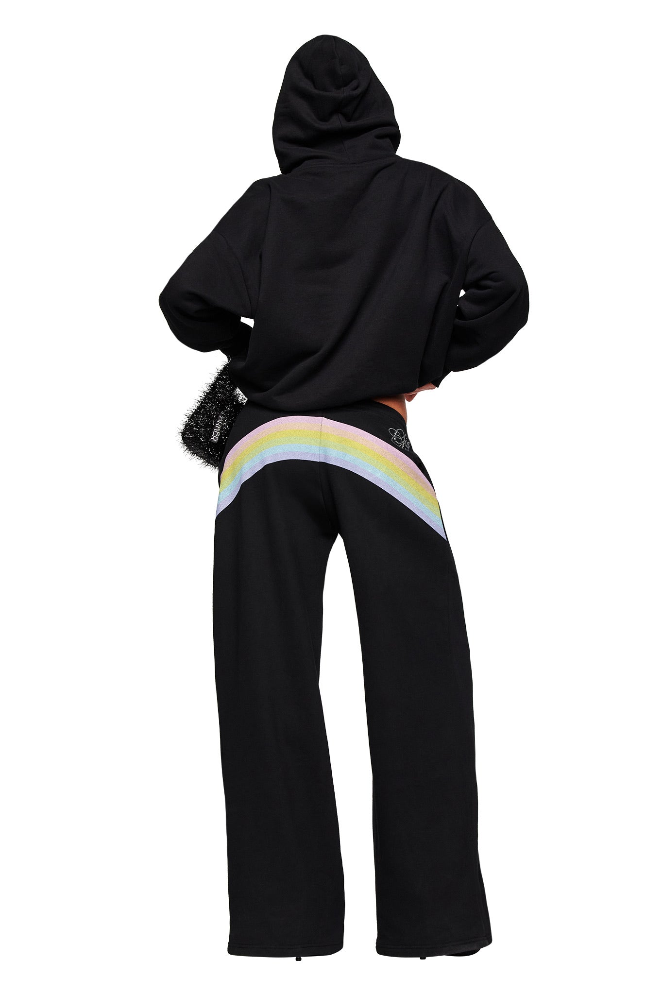 A person wears the I.AM.GIA FAFI TRACKPANT in black with a black hoodie featuring a large pastel rainbow artwork on the back, standing hands on hips and facing away from the camera against a white background.