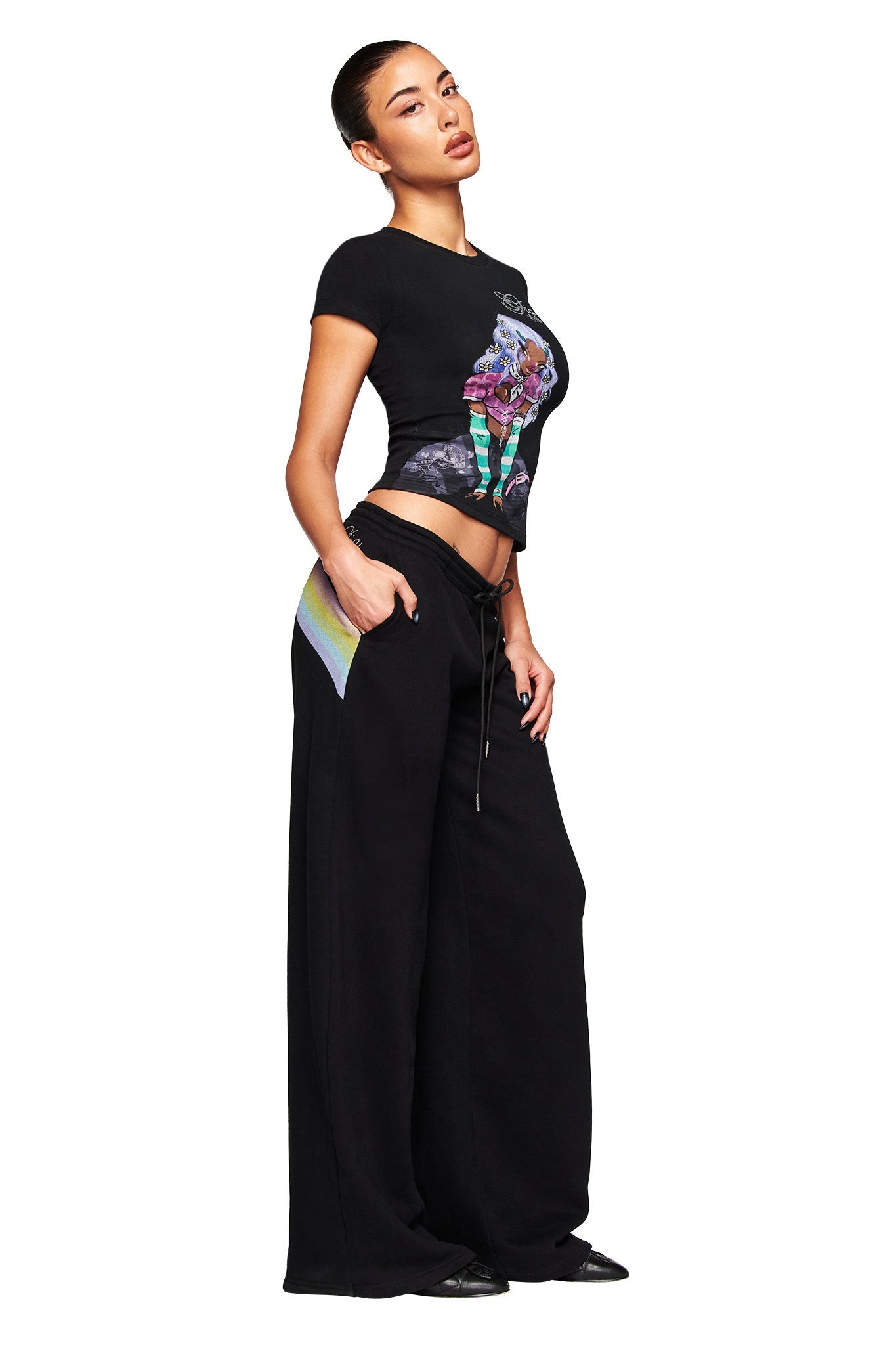 A woman poses confidently in a black graphic t-shirt and I.AM.GIA FAFI TRACKPANT - BLACK, featuring a low rise waist and pockets, paired with black shoes against a white background.