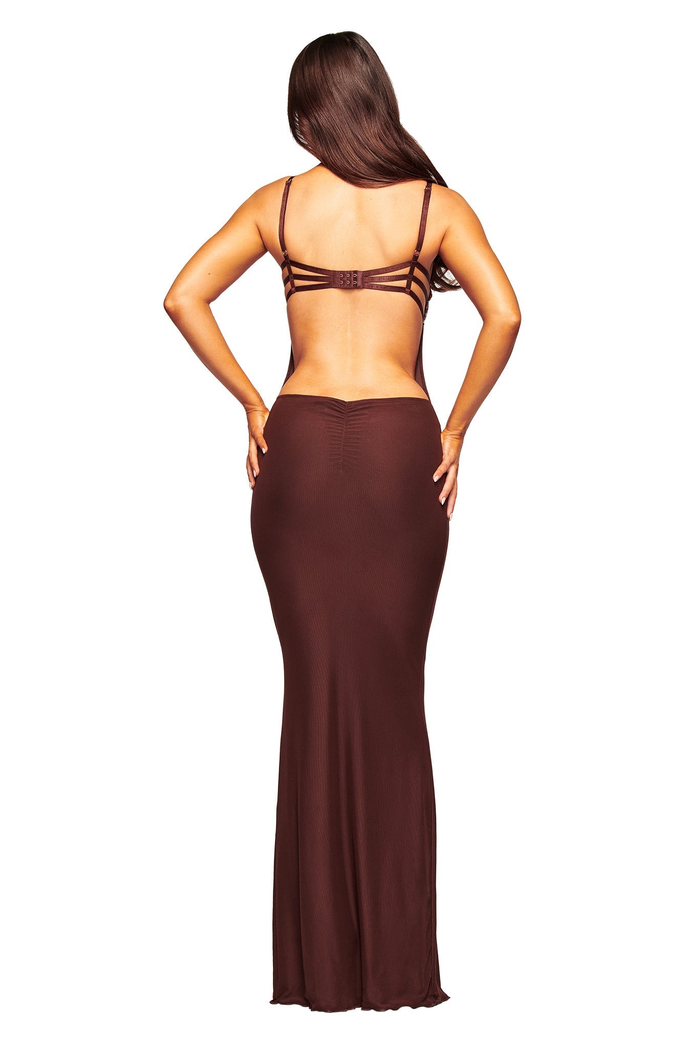 A woman with long brown hair stands facing away, wearing the I.AM.GIA SILO MAXI DRESS - BROWN, a form-fitting slip dress with thin straps, her hands resting on her hips.