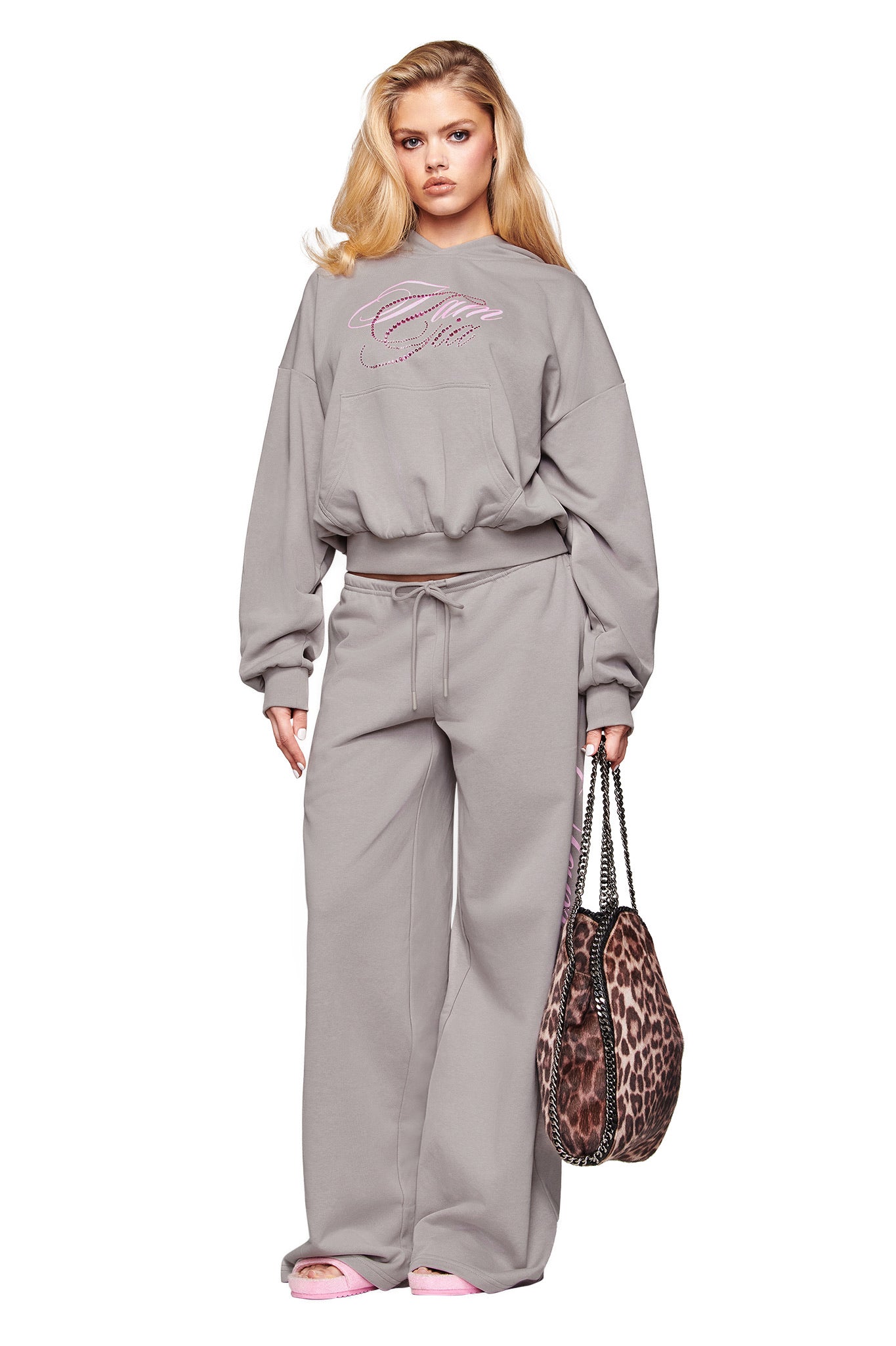 A woman wears the I.AM.GIA KADE PANT in grey, paired with a loose gray sweatshirt. She carries a leopard-print bag, sports pink slippers, and her long blonde hair falls in soft waves.