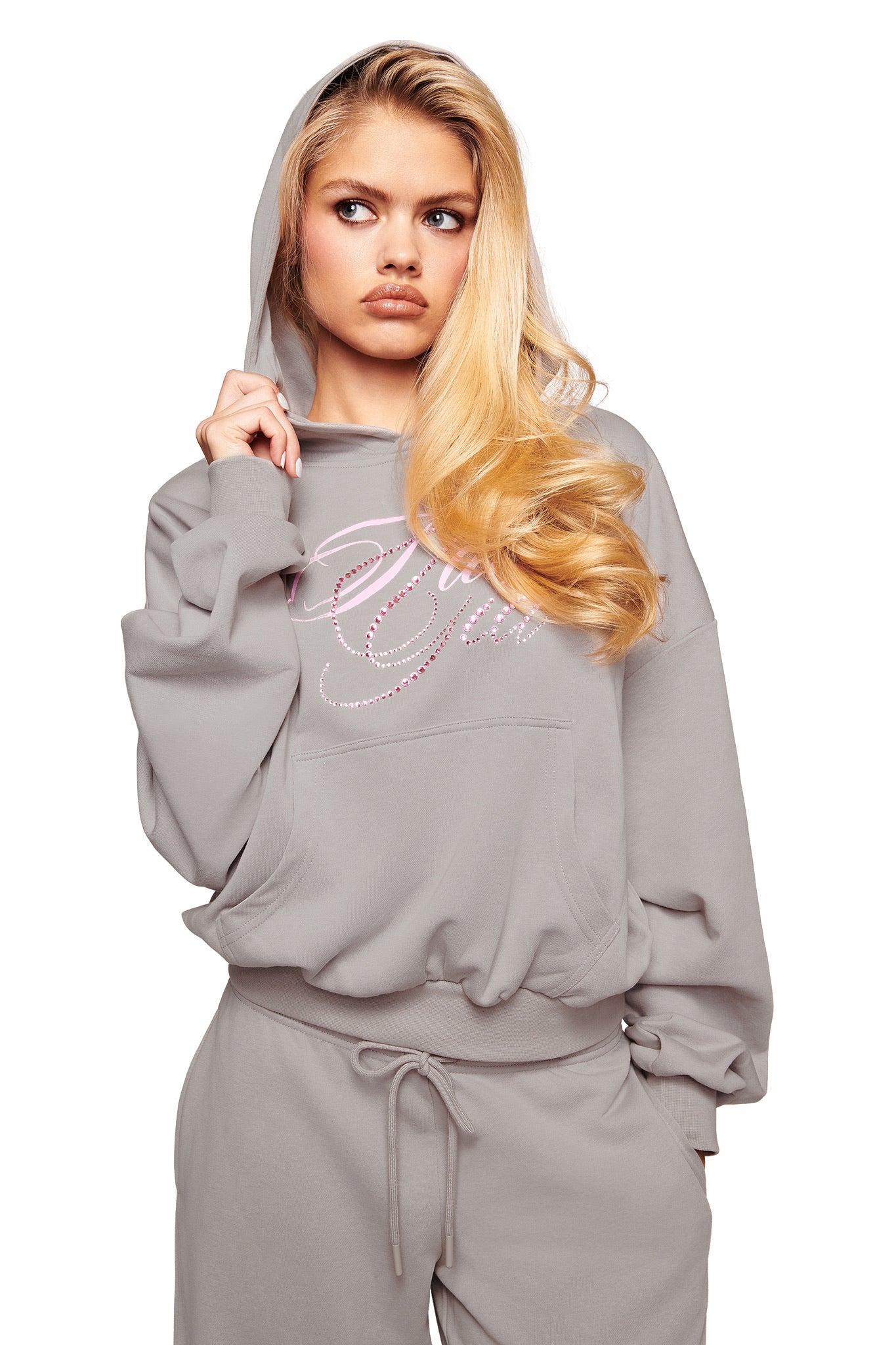 A woman with long blonde hair wears the I.AM.GIA KADE HOODIE in grey, styled cropped with the hood up and paired with matching sweatpants. She looks to the side seriously, and the Gia logo stands out against a white background.