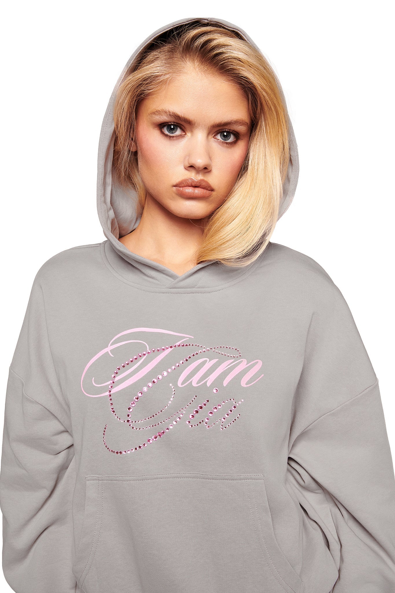 A young woman with blonde hair wears the KADE HOODIE - GREY by I.AM.GIA, a cropped light gray hoodie with the pink Gia logo in cursive on the front, standing against a white background.
