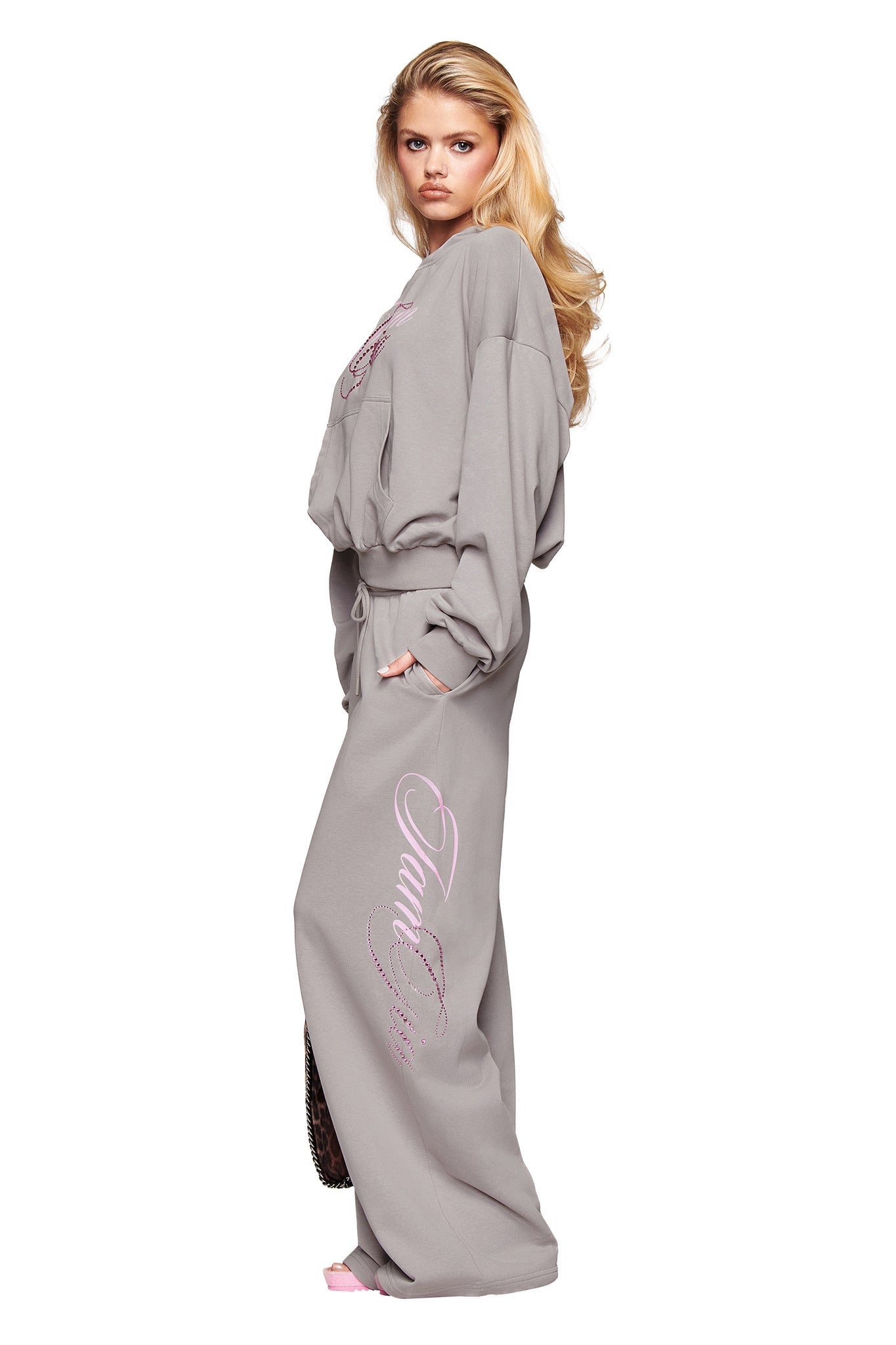 A woman with long blonde hair wears the I.AM.GIA KADE HOODIE in grey and matching sweatsuit with pink Gia logo script on the pants, standing in profile against a white background, looking seriously at the camera.