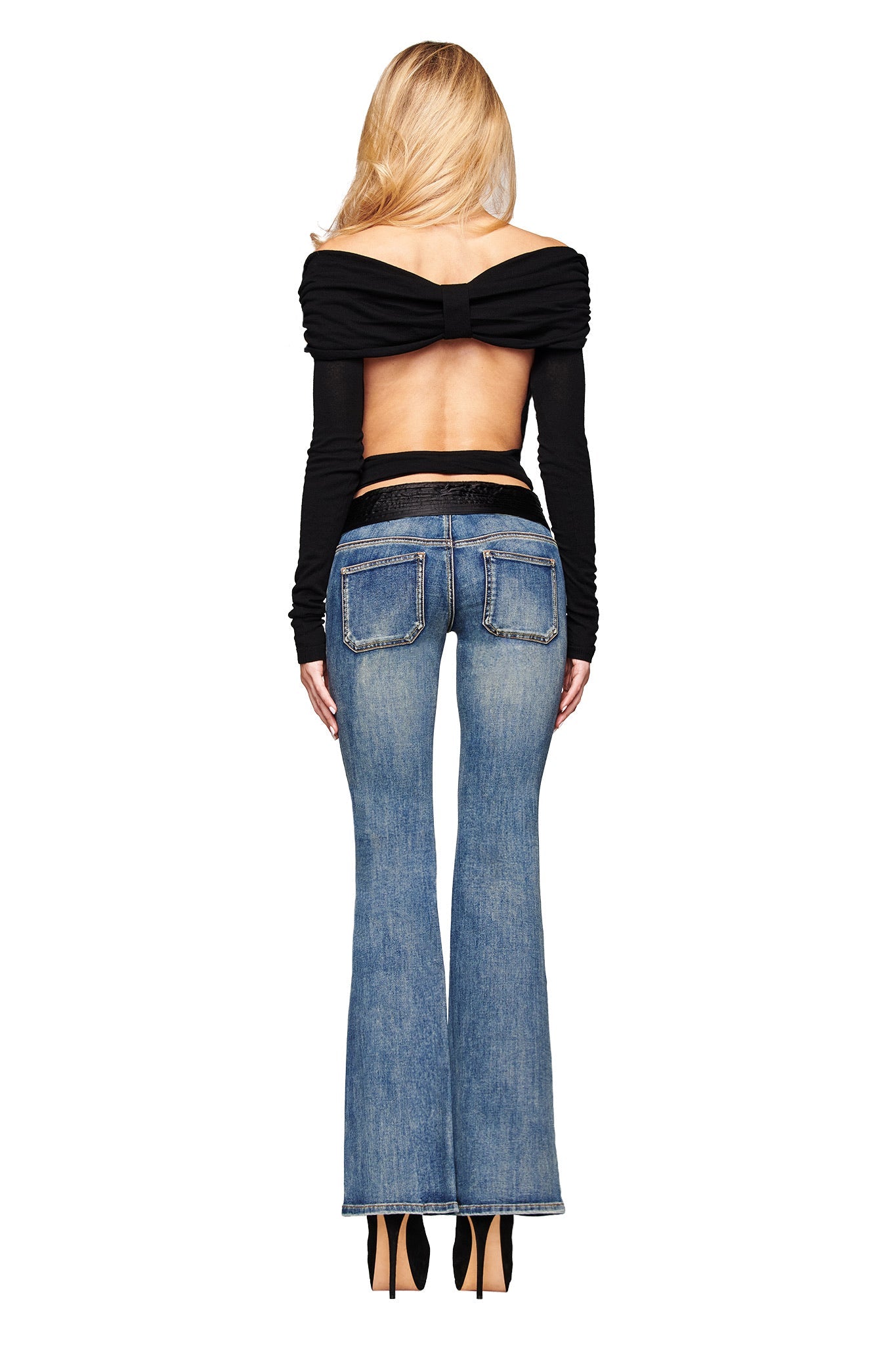 A woman with long blonde hair, facing away, wears I.AM.GIA’s KESHI JEAN in blue denim wash—a flared-leg style with satin waistband and back pockets—paired with a black off-shoulder backless top and black heels against a white background.