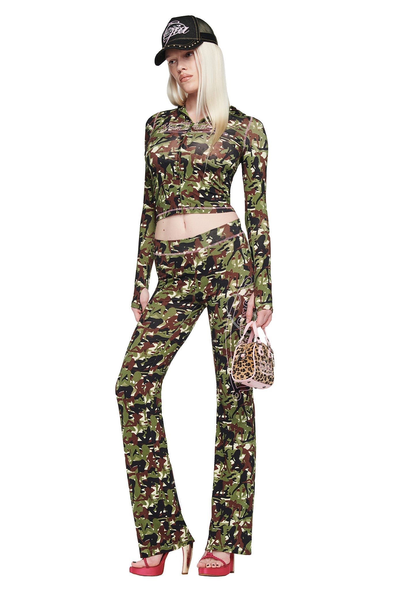 A woman wears the I.AM.GIA X VON DUTCH BLARE TRACKPANT in Stripper Camo, paired with a matching crop top, pink heels, black mesh cap, and a small leopard print handbag, standing against a white background.