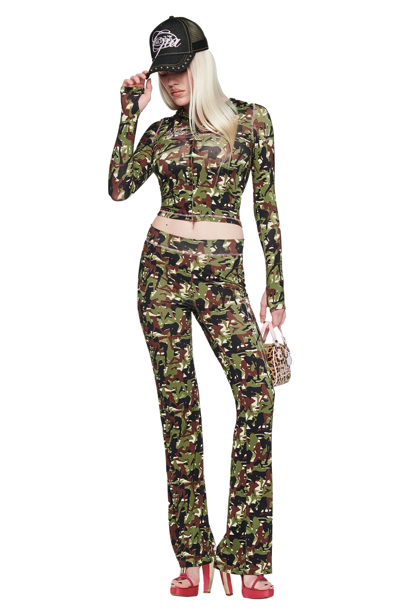 A woman with long blonde hair wears the I.AM.GIA X VON DUTCH BLARE HOODIE in camo, paired with matching pants and red platform heels. She holds a small animal-print bag and stands against a white background, channeling bold streetwear vibes.