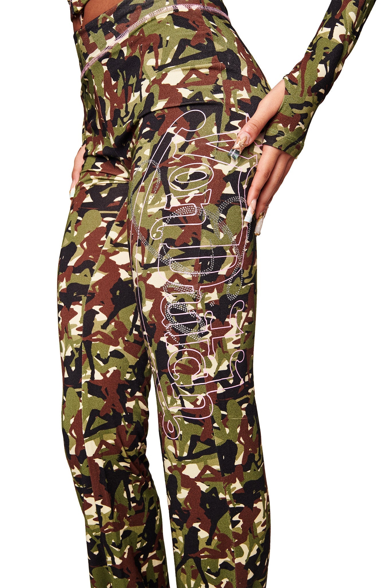 A person wears I.AM.GIA X VON DUTCH BLARE TRACKPANT - STRIPPER CAMO, featuring a camouflage pattern and white graffiti text on the upper thigh, shown waist-down with one hand on their hip.